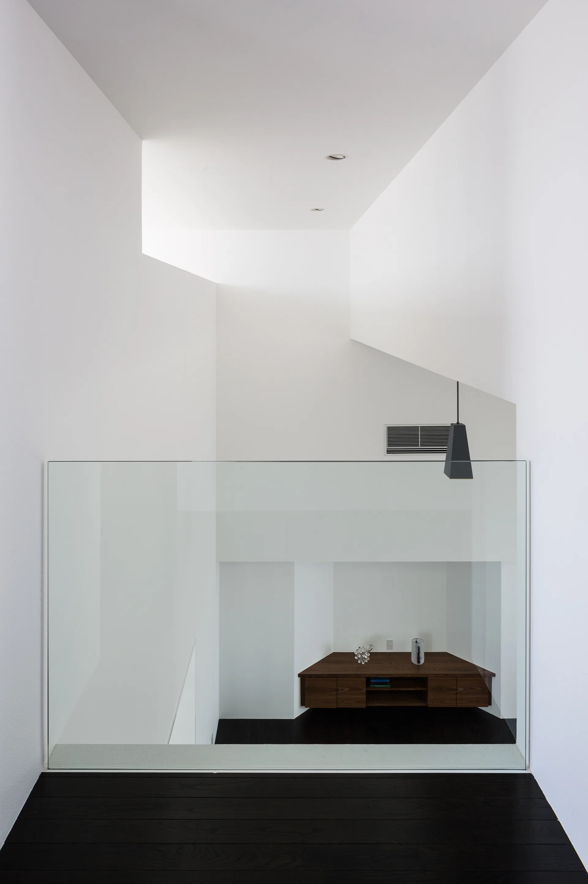 A high-angle view of a minimalist white stair landing with a glass railing looking down onto a lower level with a dark wood console table.