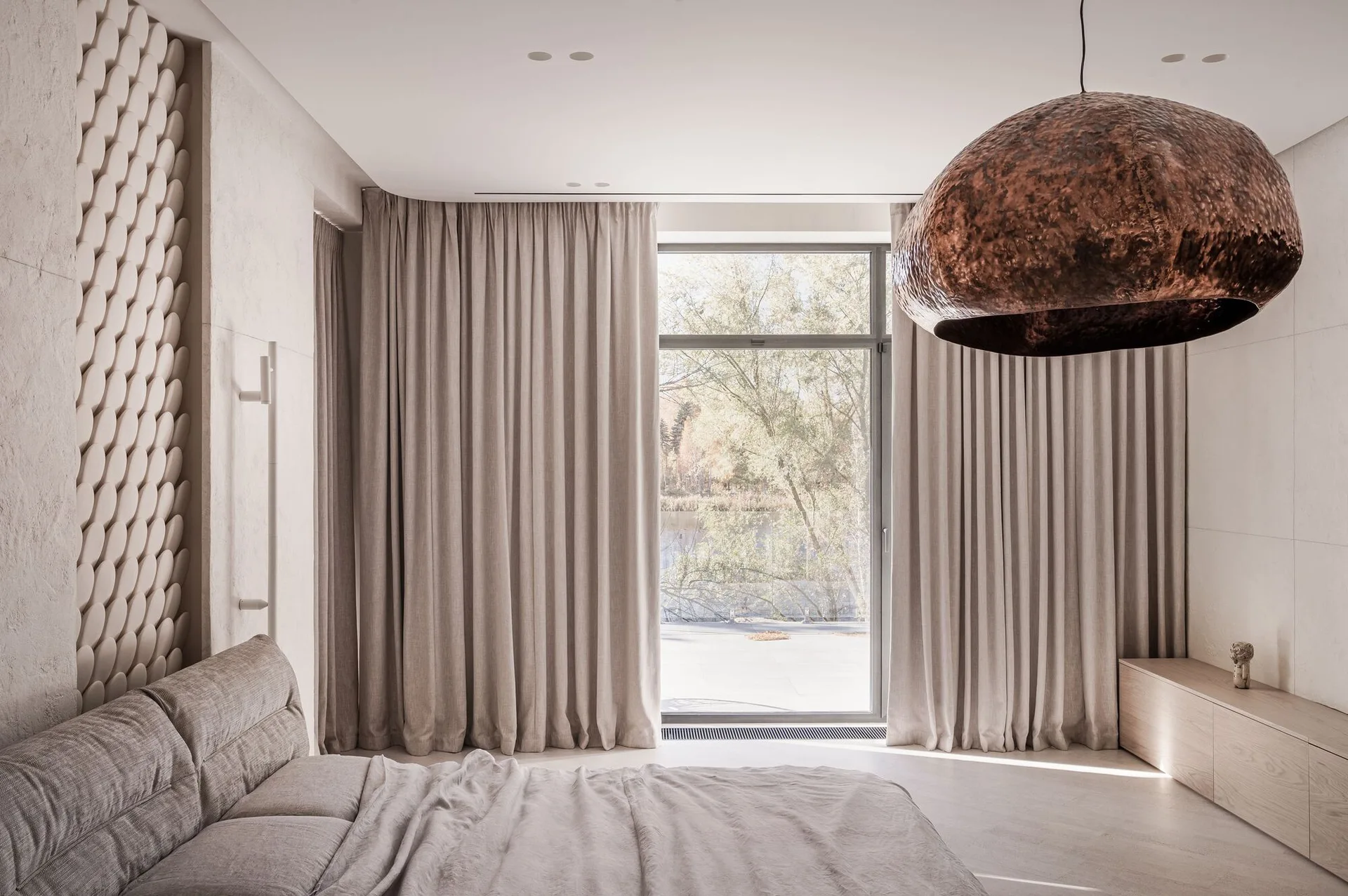 A neutral bedroom features an upholstered bed, a large hammered bronze pendant light, and floor-to-ceiling beige curtains next to a window.