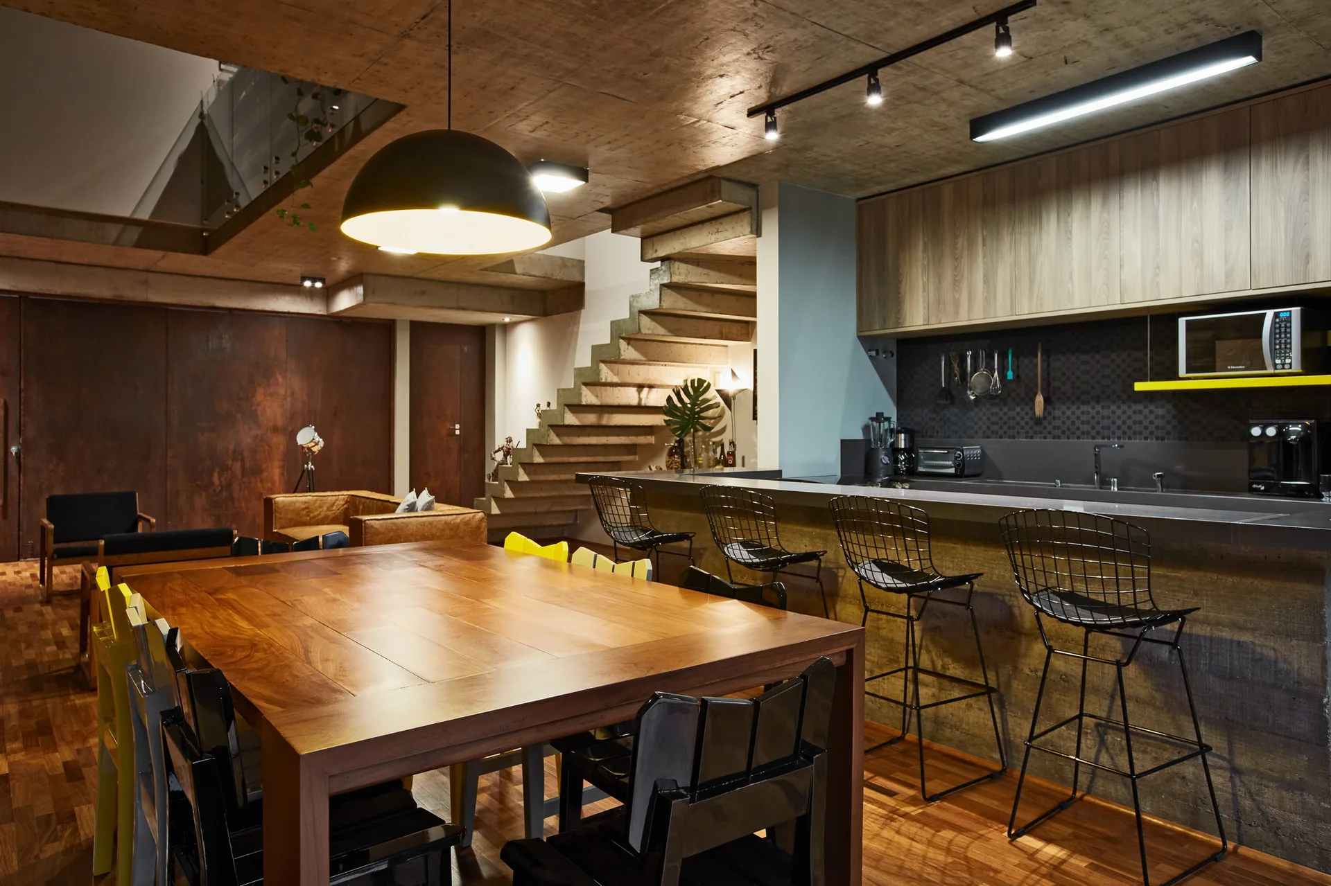 An industrial kitchen and dining space with an exposed concrete ceiling, a large wooden table, and a concrete island with wire stools.