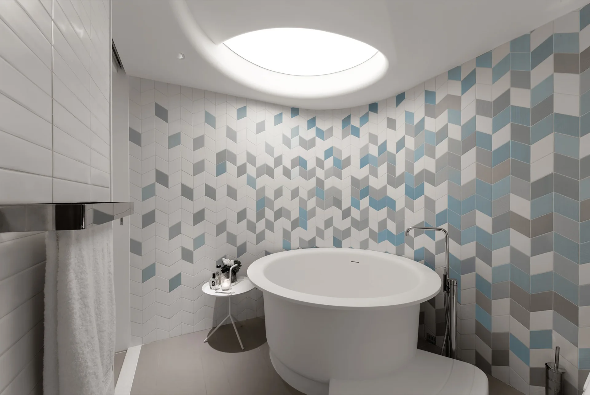 A round white freestanding bathtub sits on a light grey floor below a circular skylight and against a blue and grey chevron tiled wall.