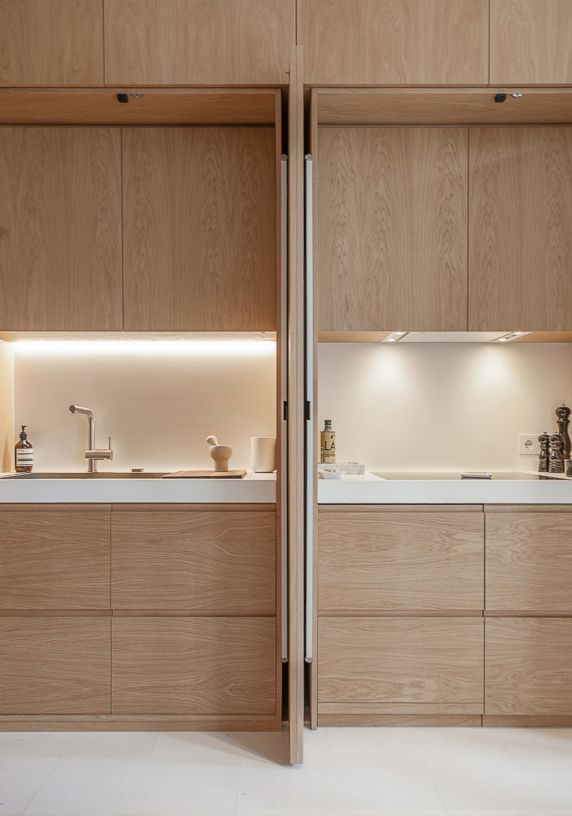 Flat-panel wood cabinets open to reveal a minimalist kitchen prep area with a white countertop, faucet, cooktop, and warm under-cabinet lighting.