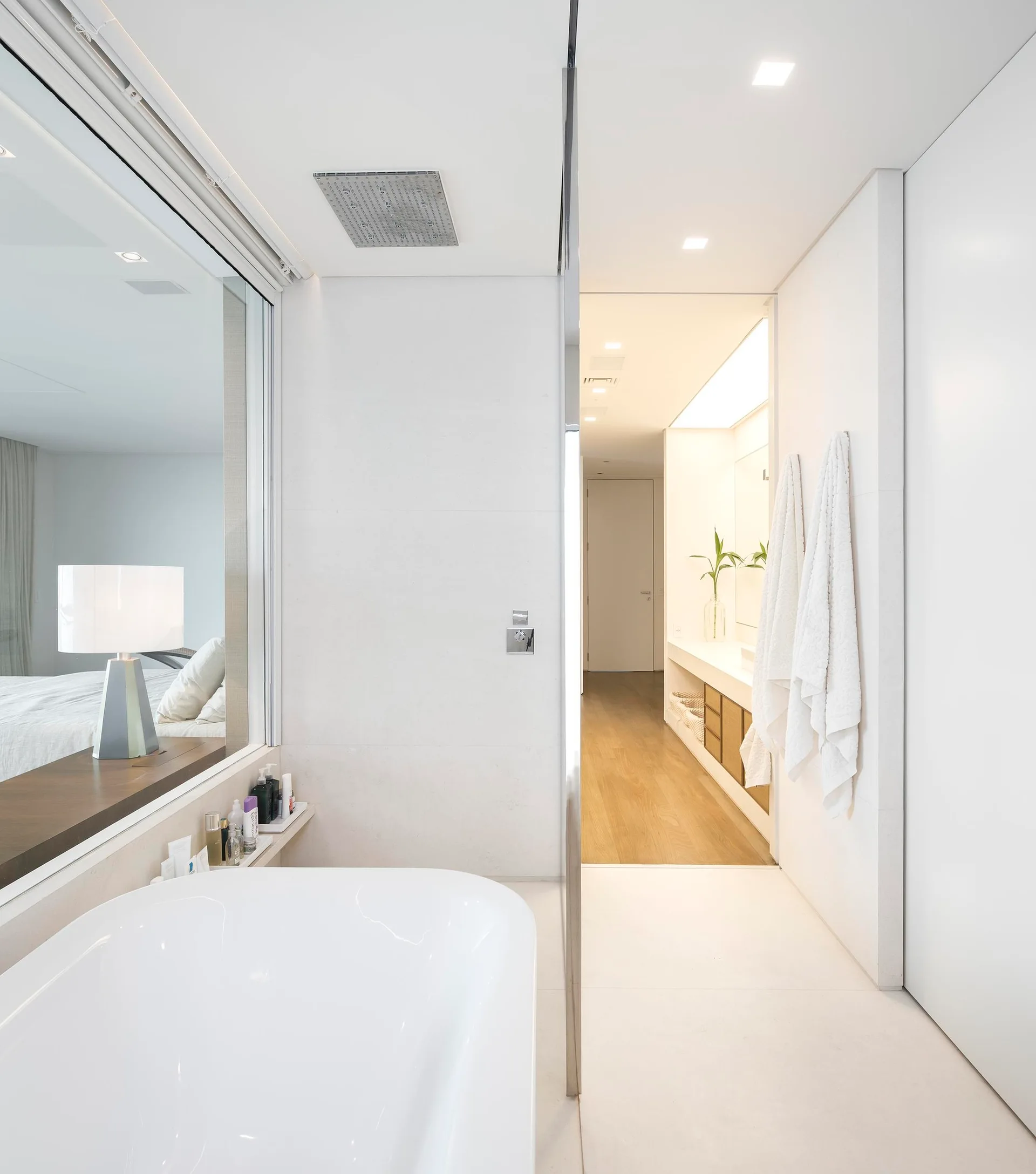 A minimalist white bathroom features a soaking tub, a ceiling-mounted shower head, and a large glass partition looking into an adjacent bedroom.