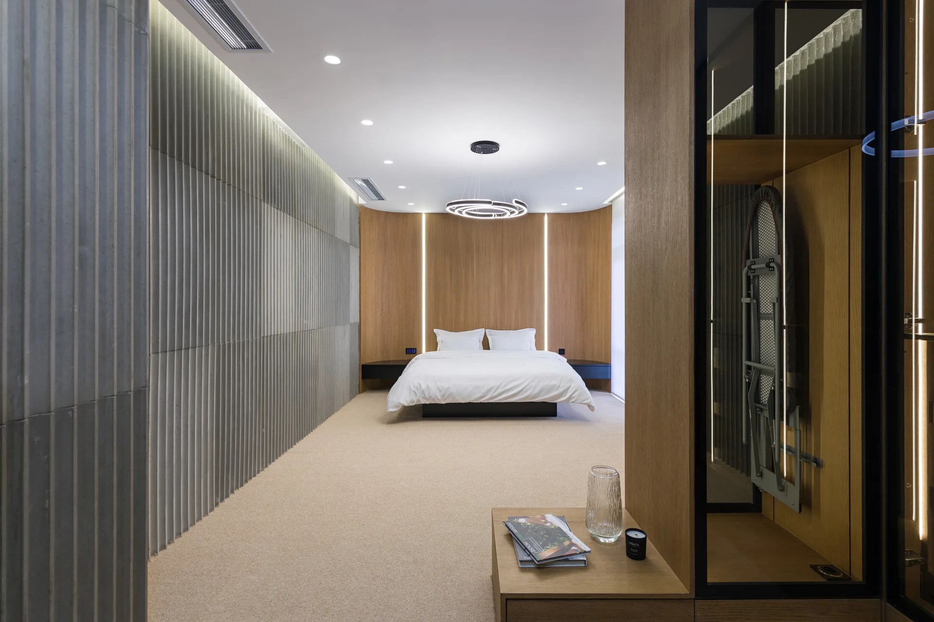A minimalist bedroom featuring a bed set against a curved wood-paneled wall, alongside vertical ribbed grey wall panels and warm recessed lighting.