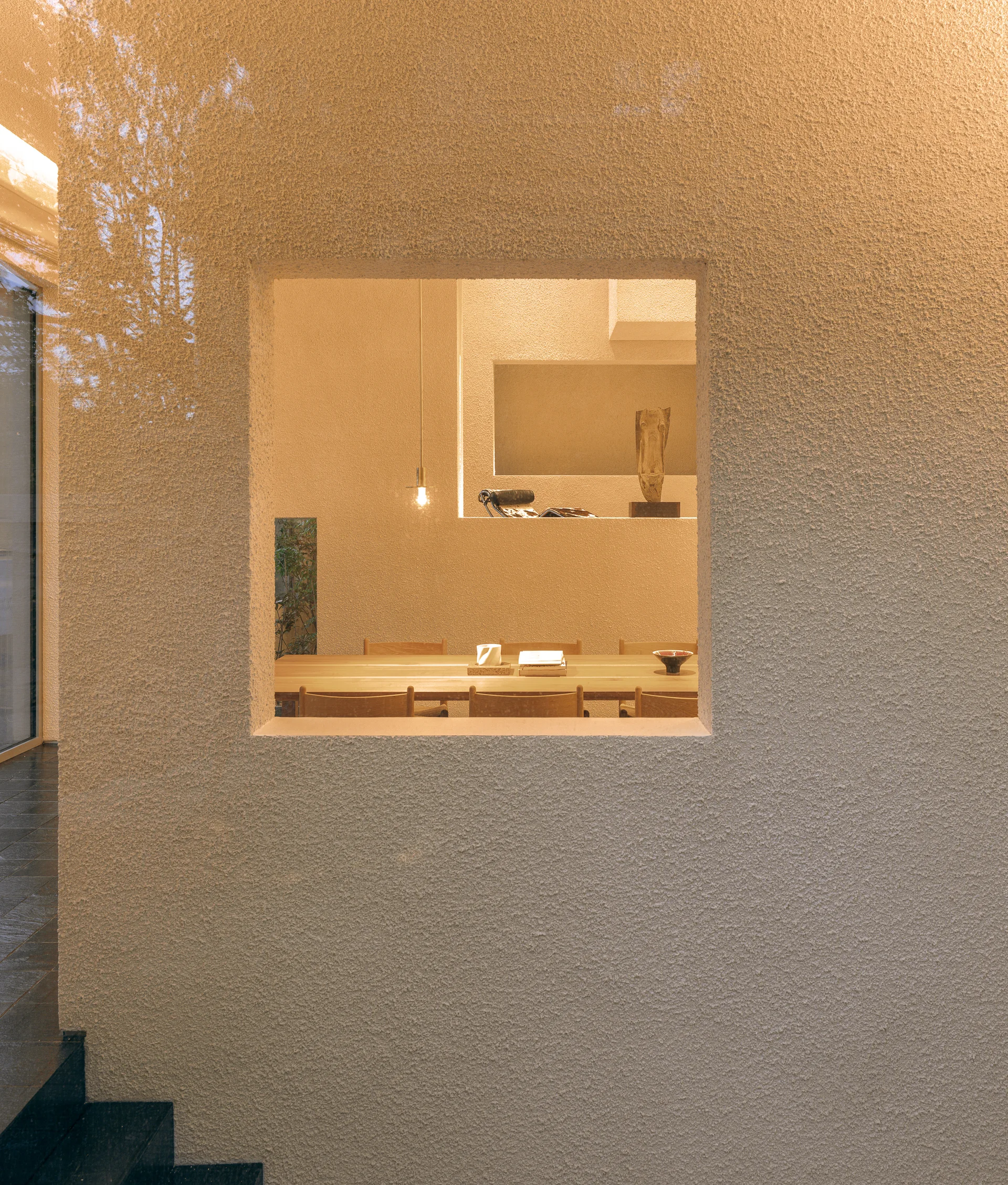A view through a square wall cutout reveals a minimalist dining room with a light wood table, warm pendant lighting, and textured beige plaster walls.