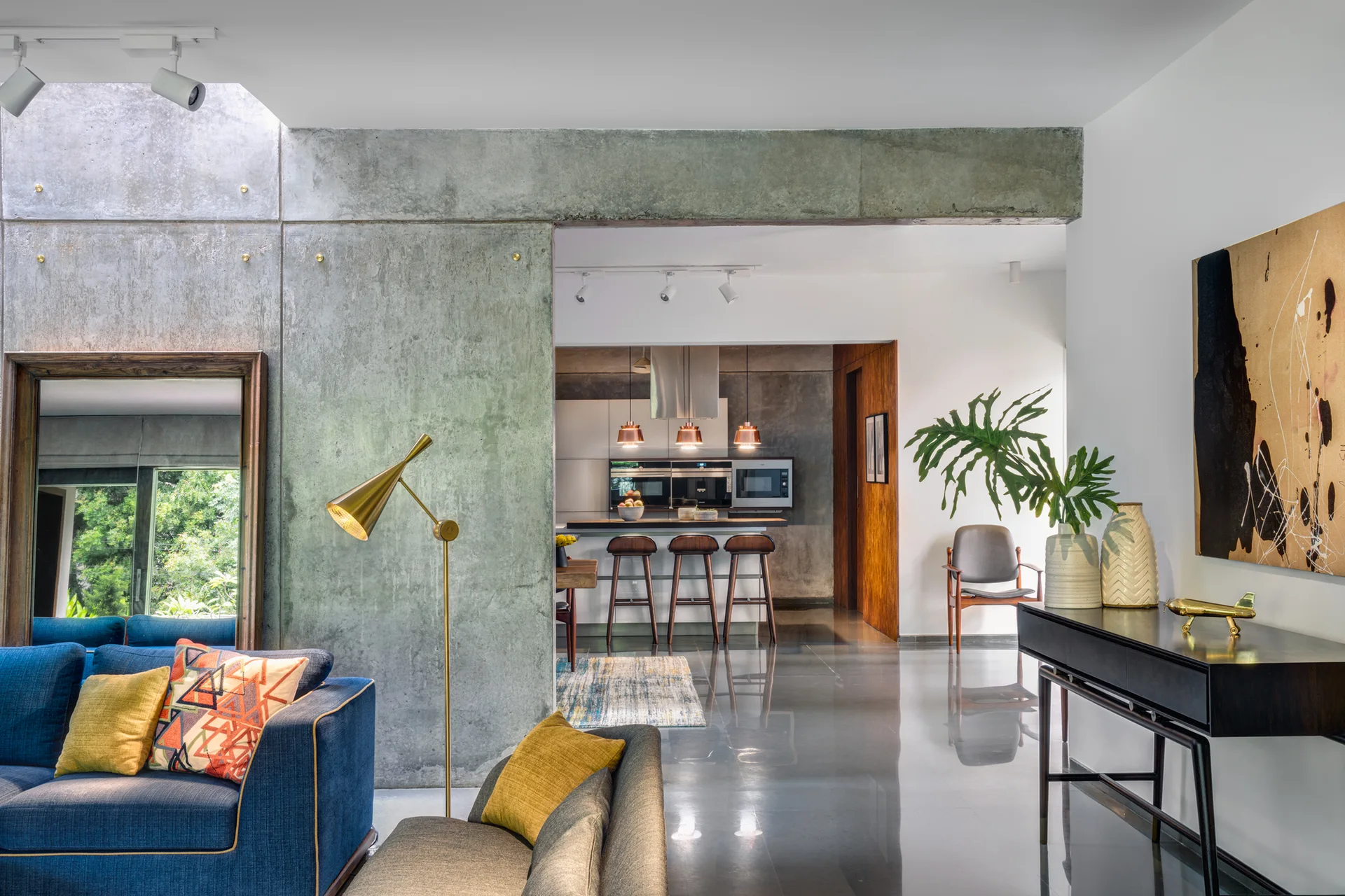 A modern open-plan living room featuring a blue sofa, a brass floor lamp, a raw concrete partition wall, and a kitchen visible in the background.