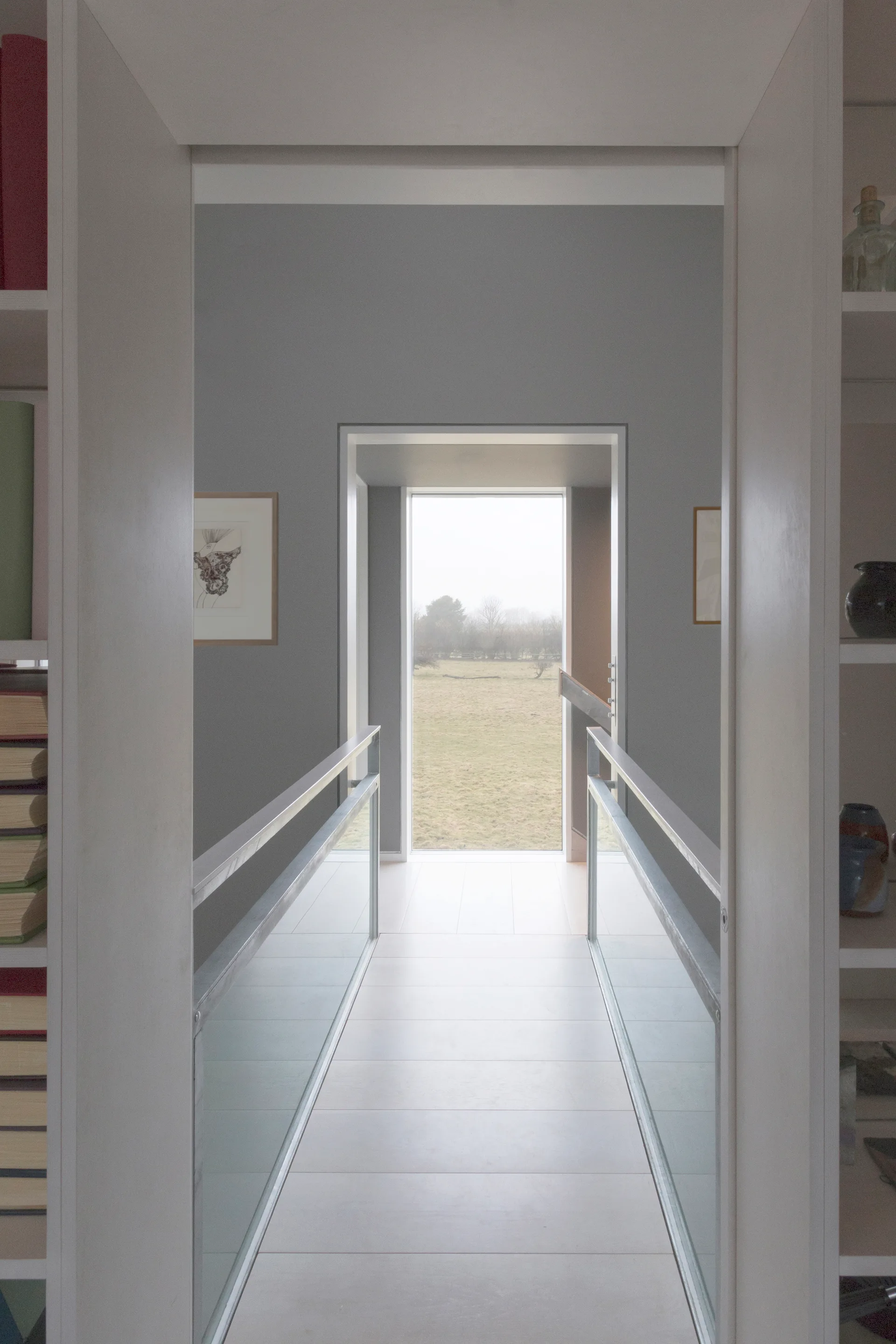 A minimalist gray hallway features white built-in bookshelves at the entrance and a glass-railed bridge leading toward a large window with a landscape view.