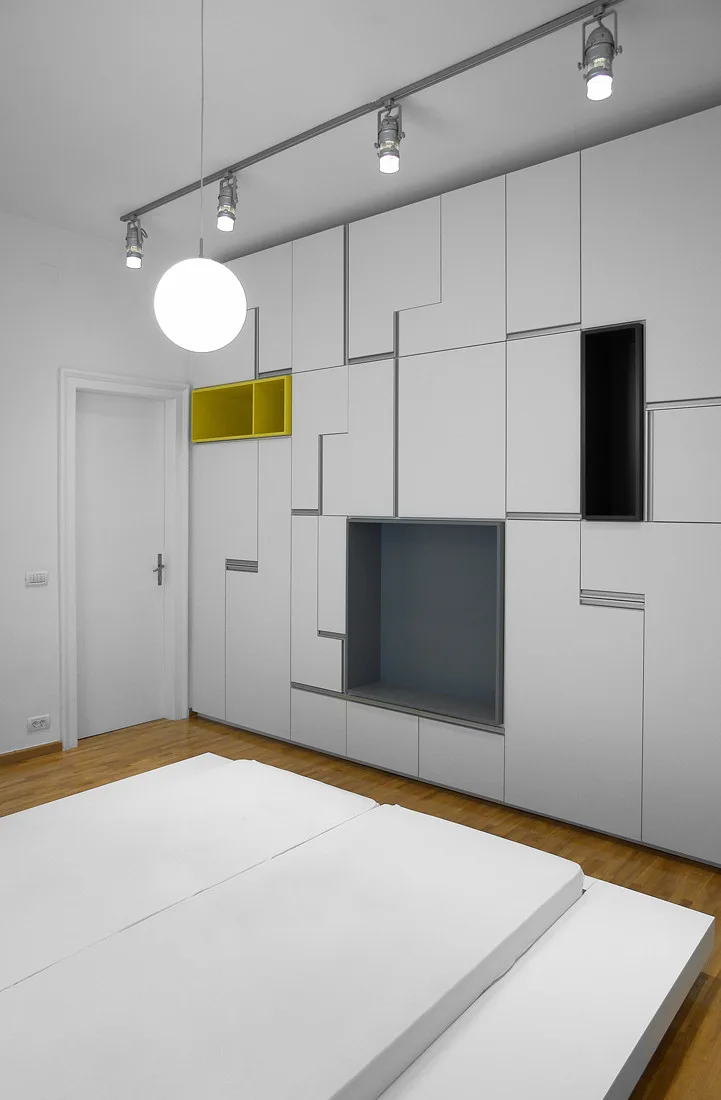 A minimalist bedroom featuring a massive white modular storage wall with recessed yellow, grey, and black niches next to low platform beds.