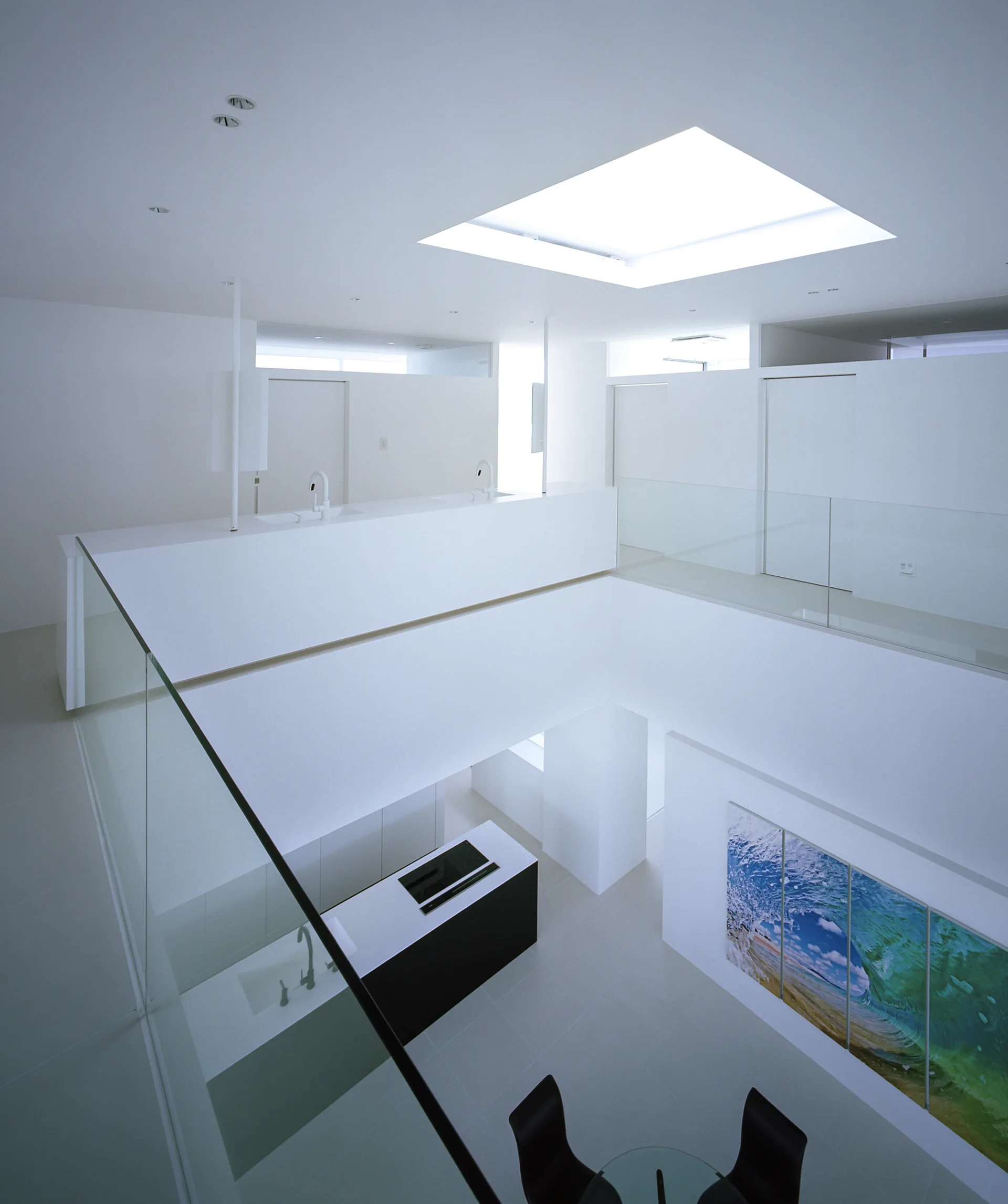 A high-angle view of a double-height white minimalist interior featuring a glass-railed mezzanine, skylight, and kitchen below.