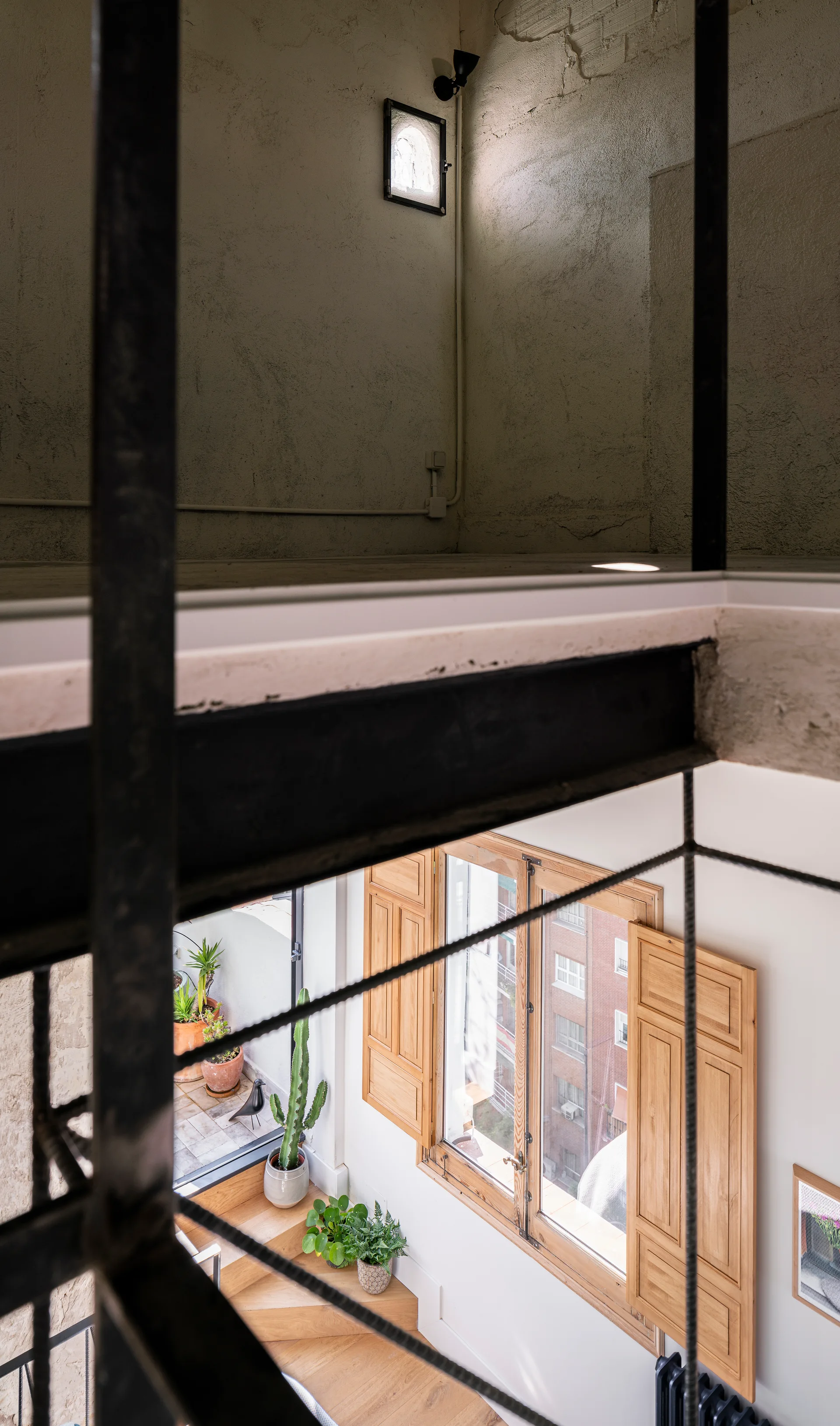 High-angle view of an industrial stairwell featuring textured plaster walls, a black steel beam, wooden stairs, and a window with open wood shutters.