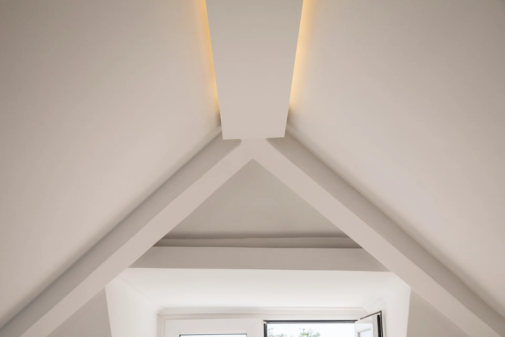 Close-up of a white vaulted ceiling with angled structural beams meeting at a ridge featuring warm recessed cove lighting.
