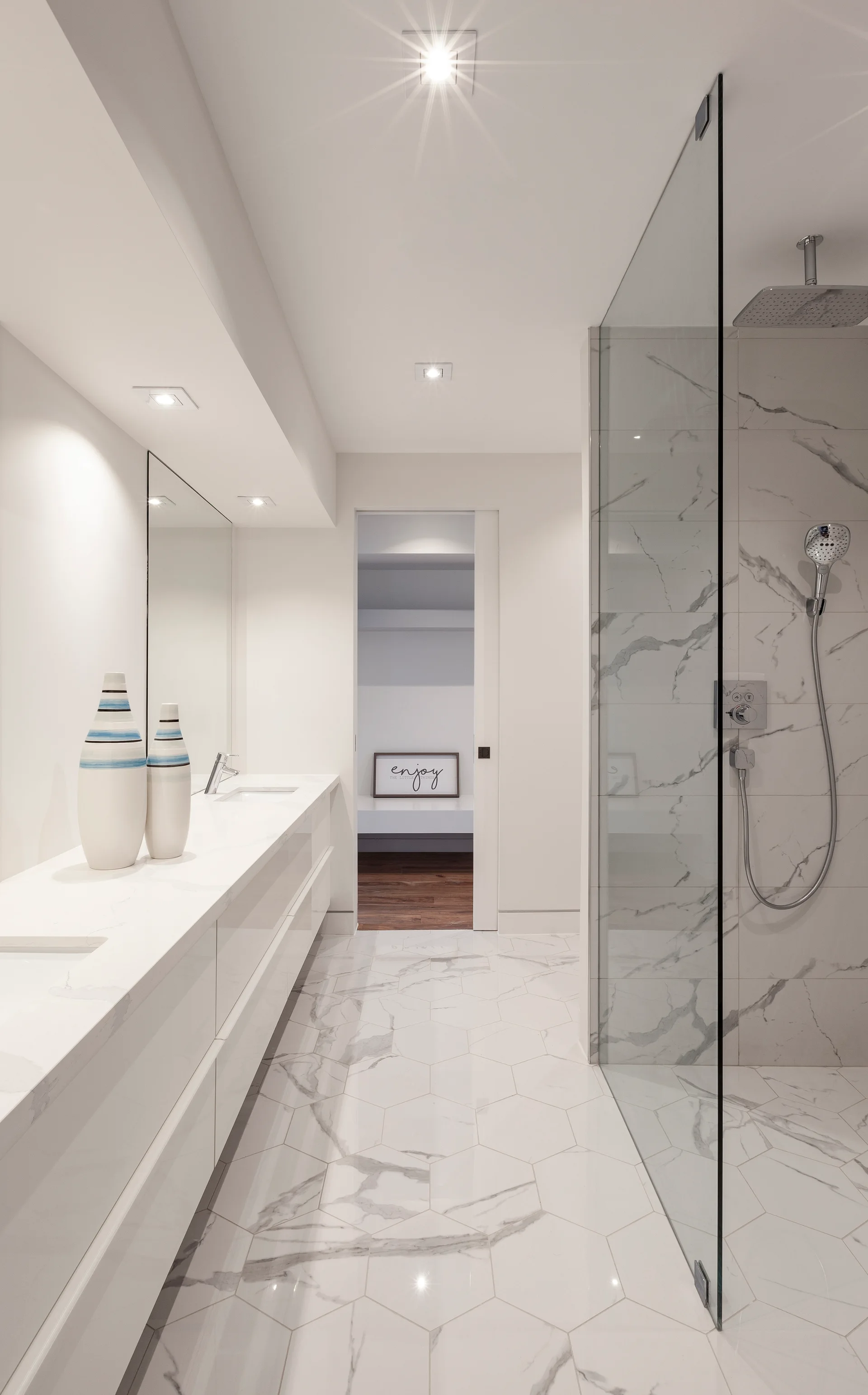 A contemporary white bathroom features a long glossy double vanity, hexagonal marble-look floor tiles, and a frameless glass walk-in shower.