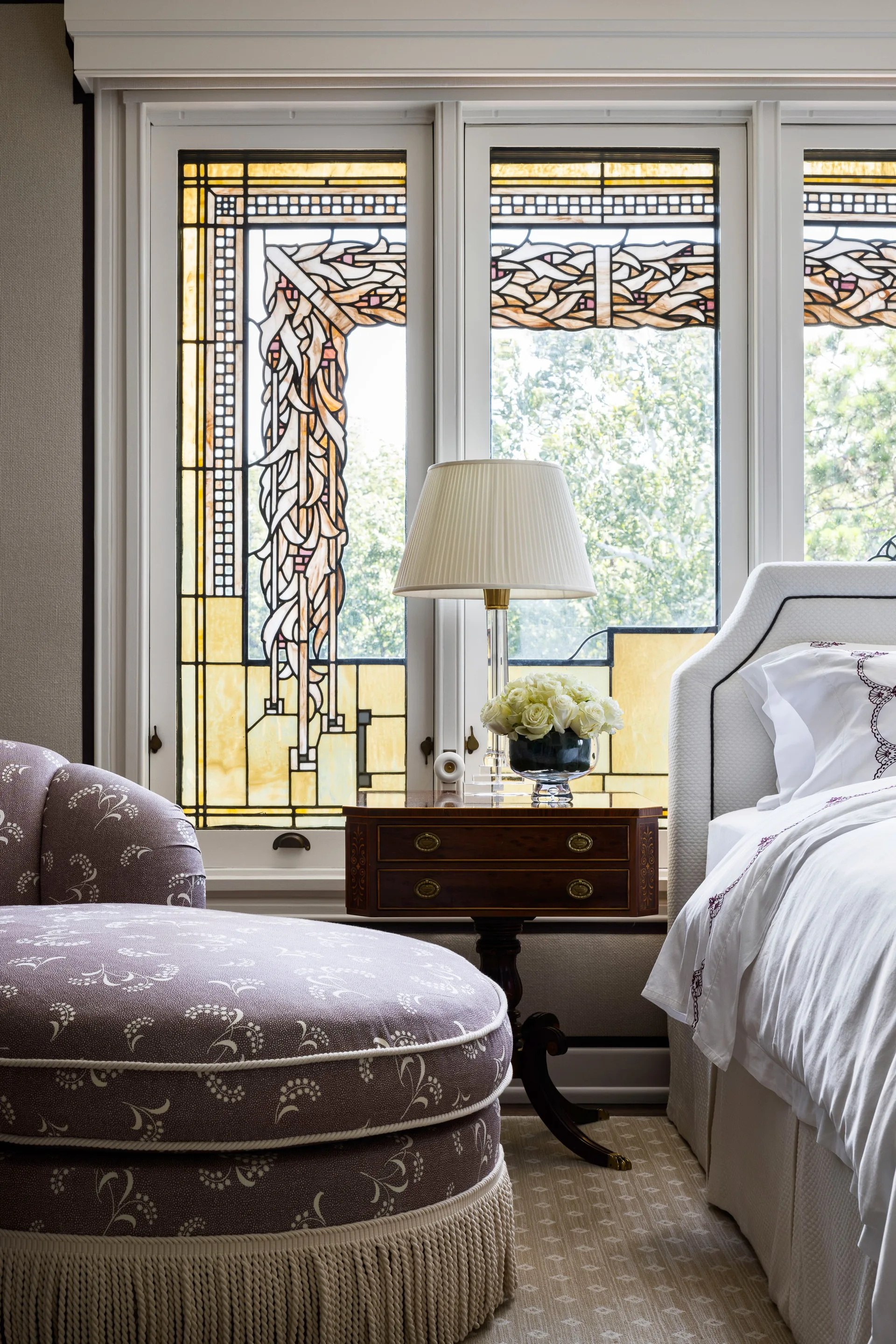 A classic bedroom featuring an upholstered purple floral chaise, a dark wood nightstand, and a leaded stained glass window.