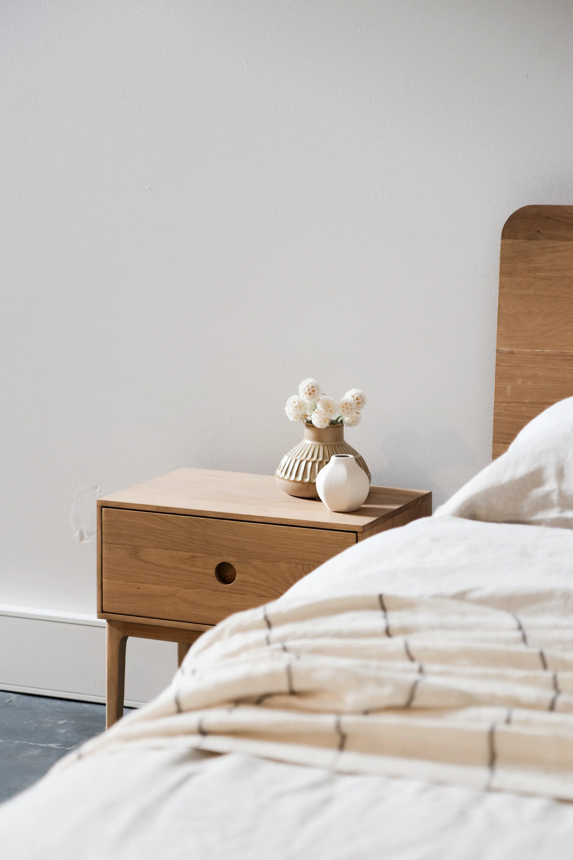 A minimalist wooden nightstand with a circular cutout drawer pull sits next to a bed dressed in checked linen bedding and a wooden headboard.