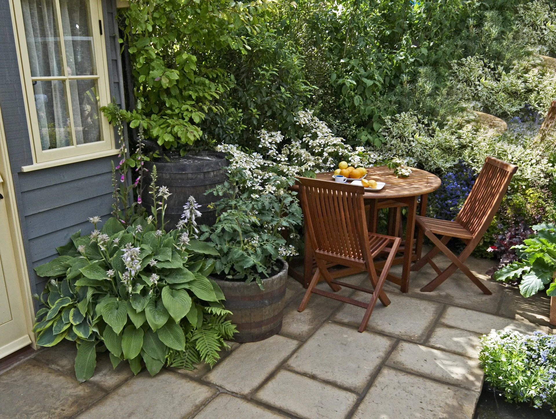 A wooden bistro table and two folding chairs sit on a stone patio surrounded by lush garden plants and a grey wooden garden shed.