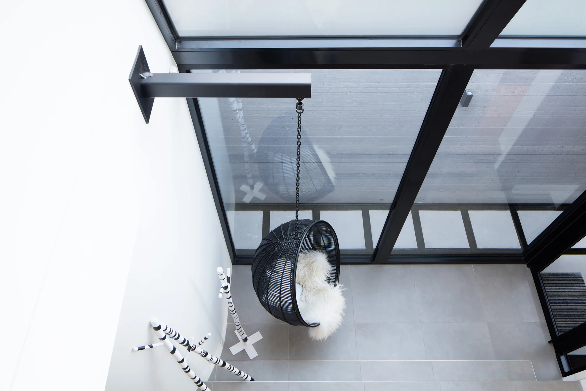 High-angle view of a black hanging egg chair with a white faux fur throw on a grey tiled floor next to a glass wall.
