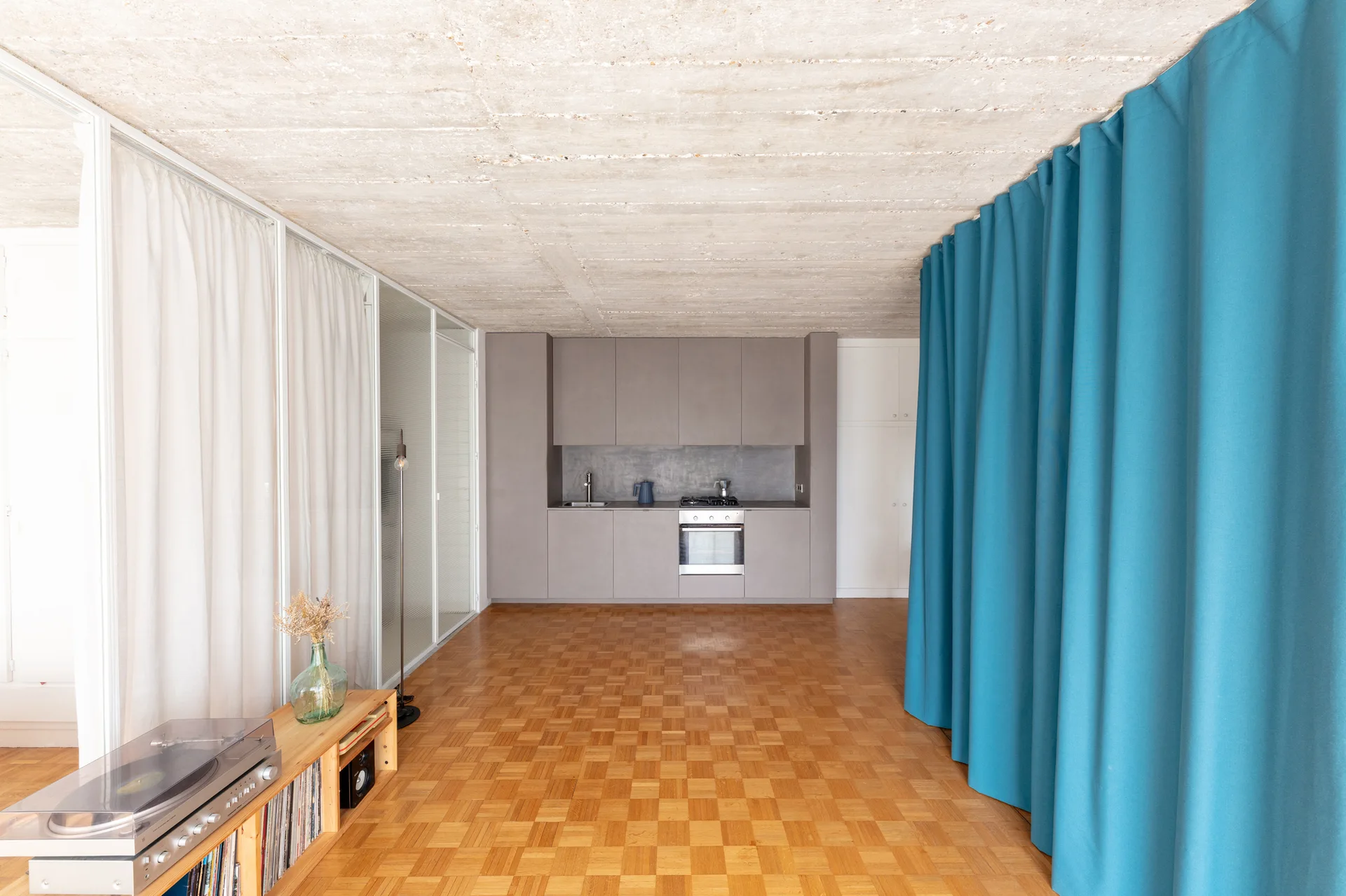 A minimalist kitchen featuring grey cabinetry, an exposed concrete ceiling, parquet flooring, and a floor-to-ceiling blue curtain partition.