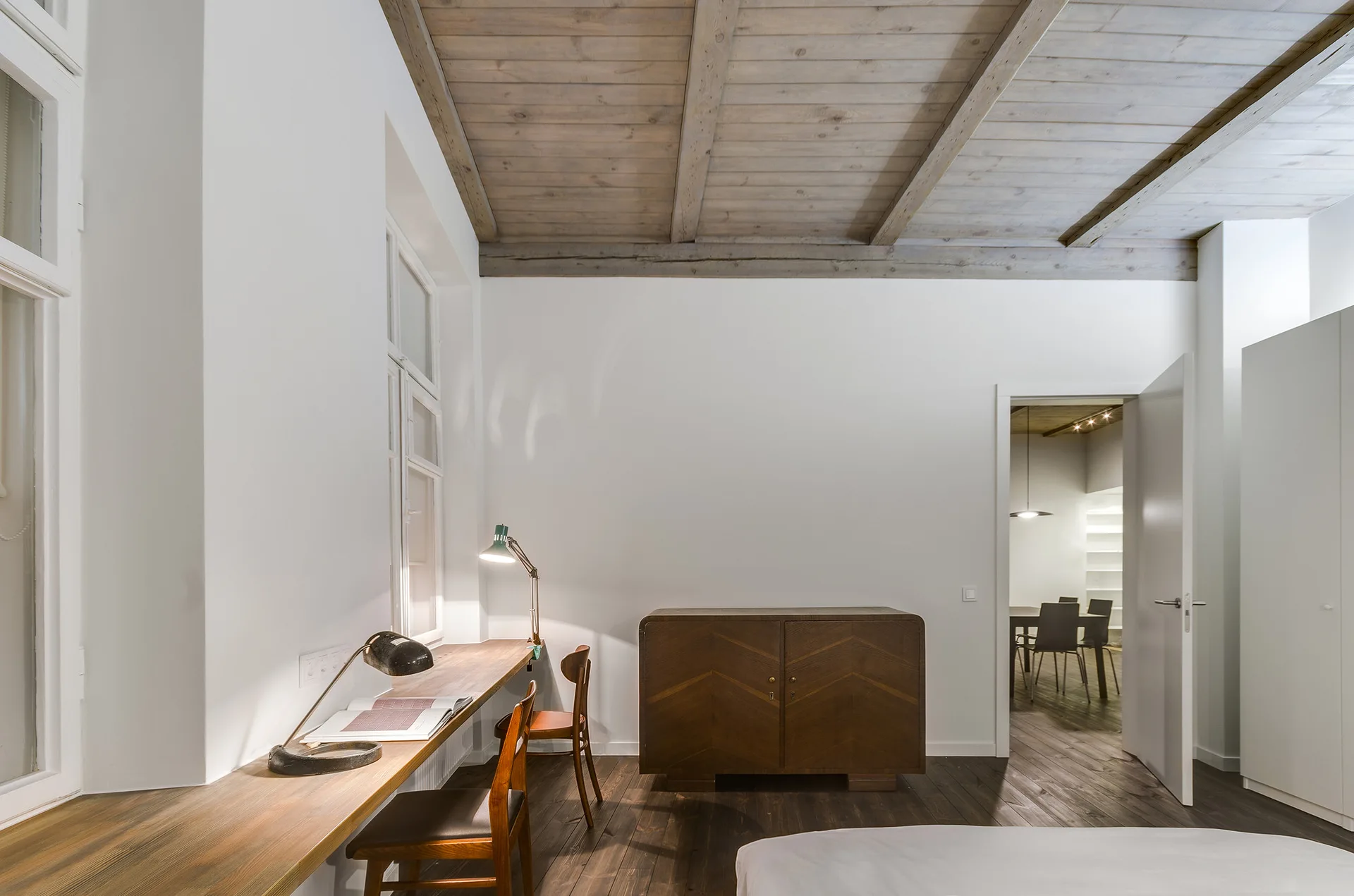 A minimalist bedroom features white walls, exposed wood ceiling beams, a long wooden built-in desk under a window, and a dark wood cabinet.