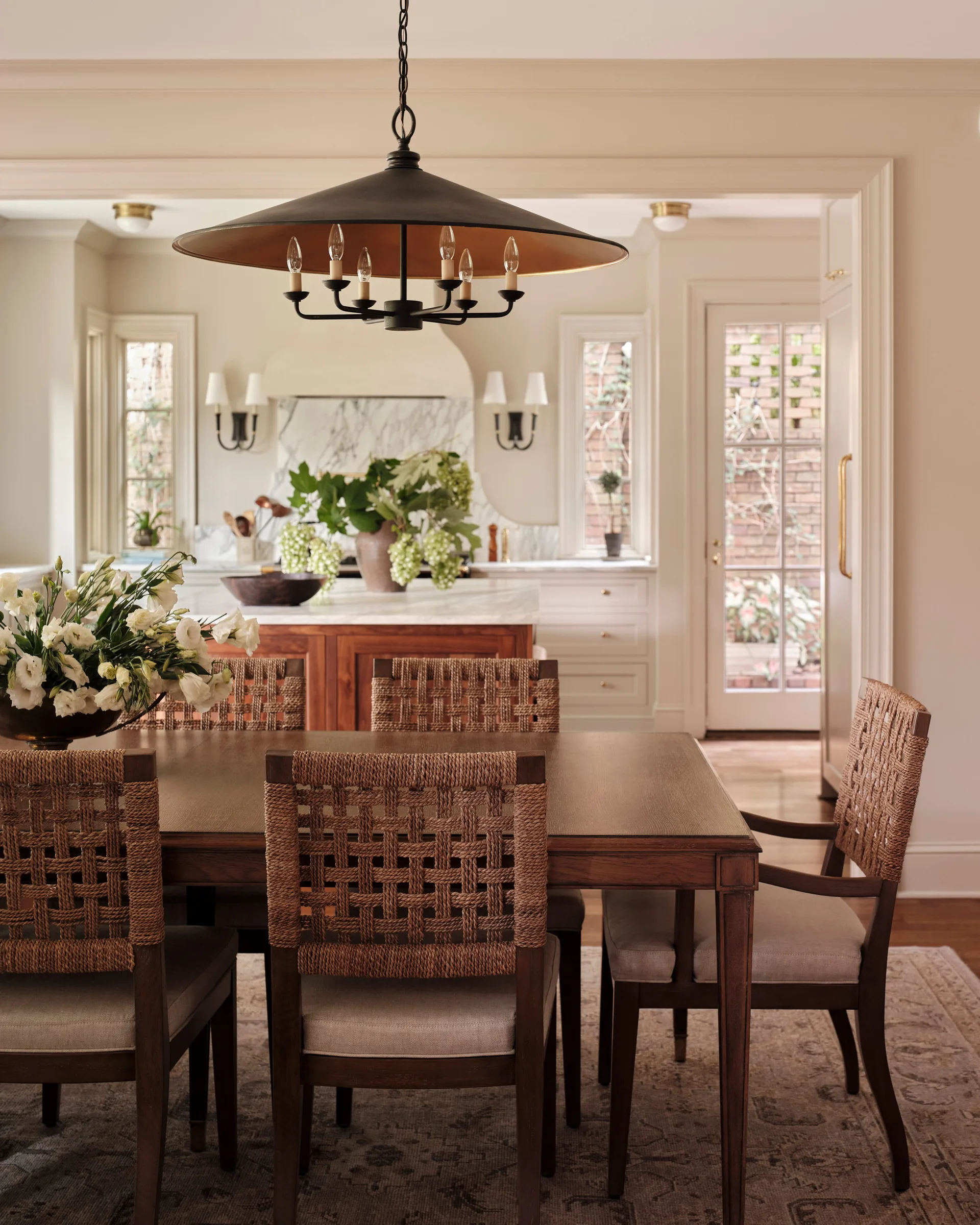 A wooden dining table surrounded by woven rope chairs sits beneath a large black pendant light in front of a bright kitchen.