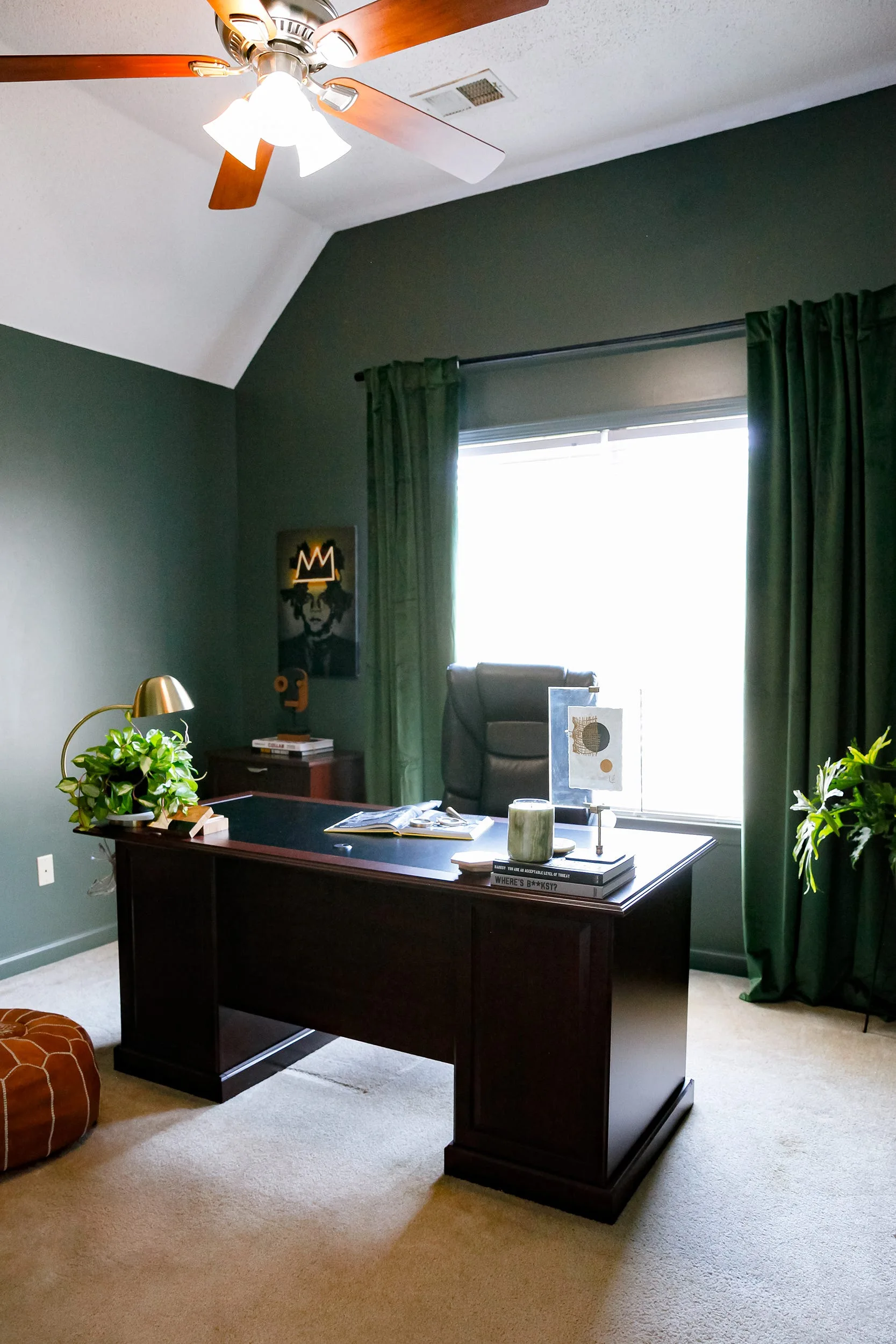 A home office features a dark wood desk, a leather office chair, deep green painted walls, and matching green velvet window curtains.