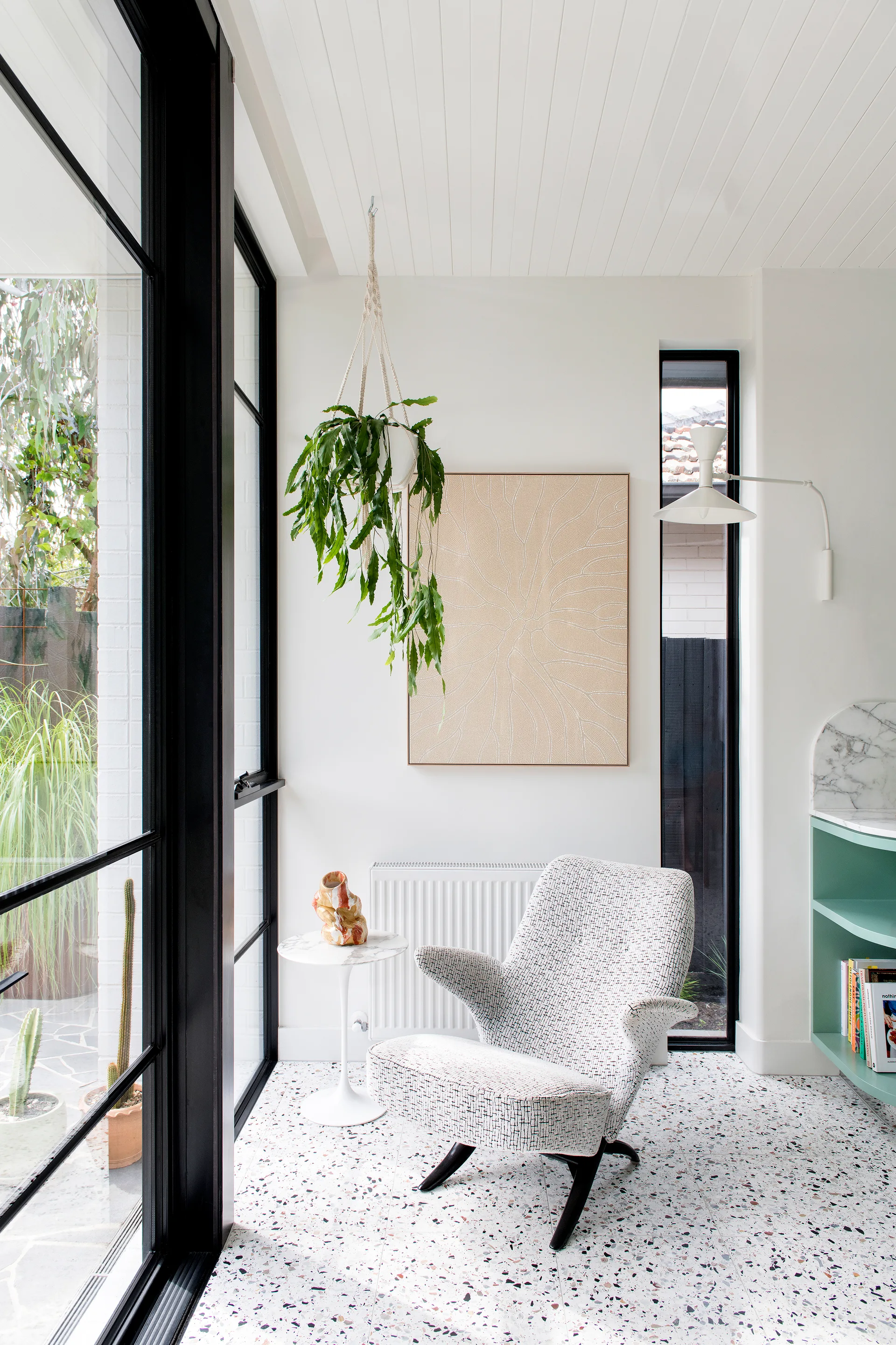 A modern sitting corner features a patterned armchair and marble side table on speckled terrazzo flooring next to large black-framed windows.