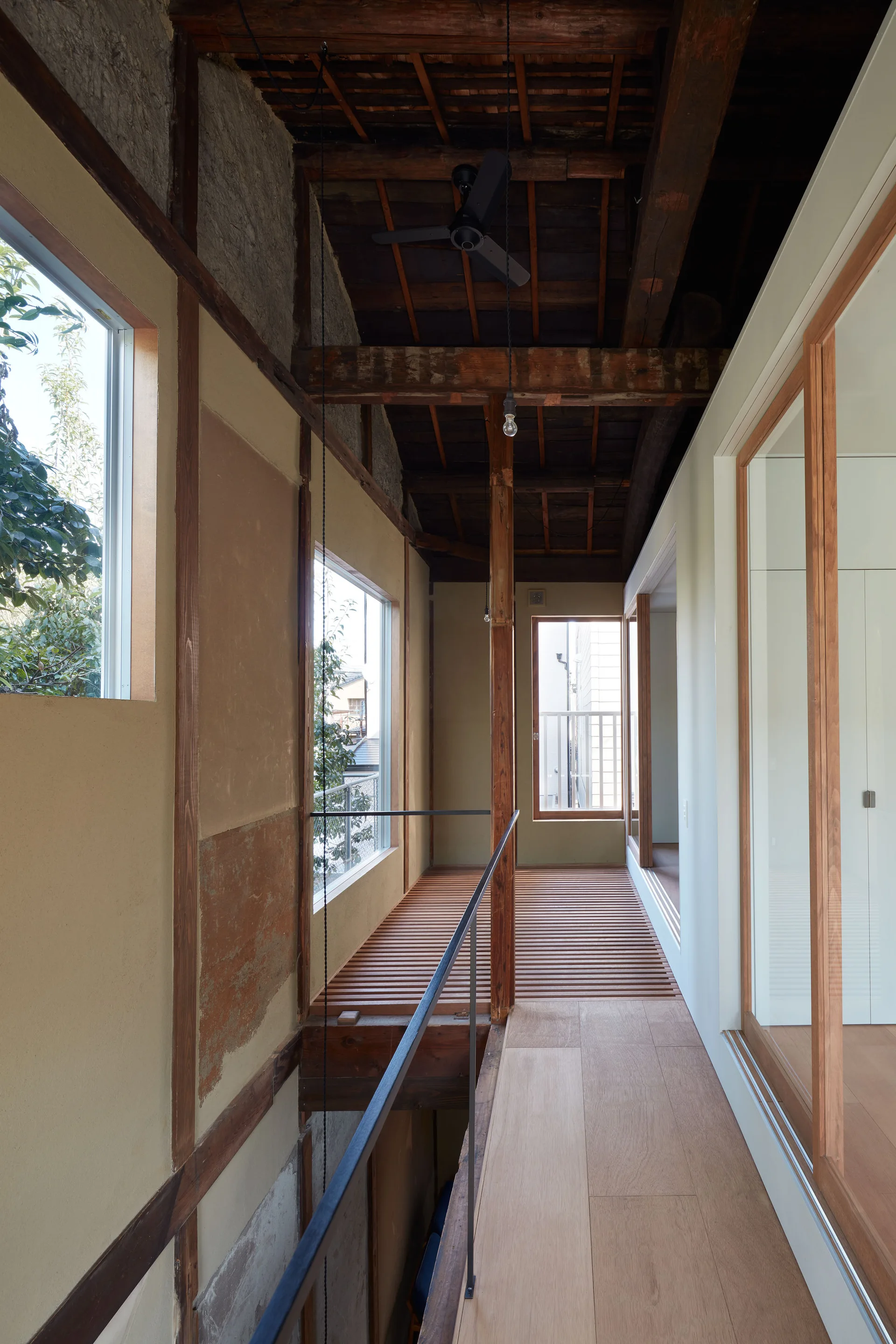 A minimalist hallway features a slatted wooden walkway, rough plaster walls, exposed dark timber ceiling beams, and a slim black metal railing.