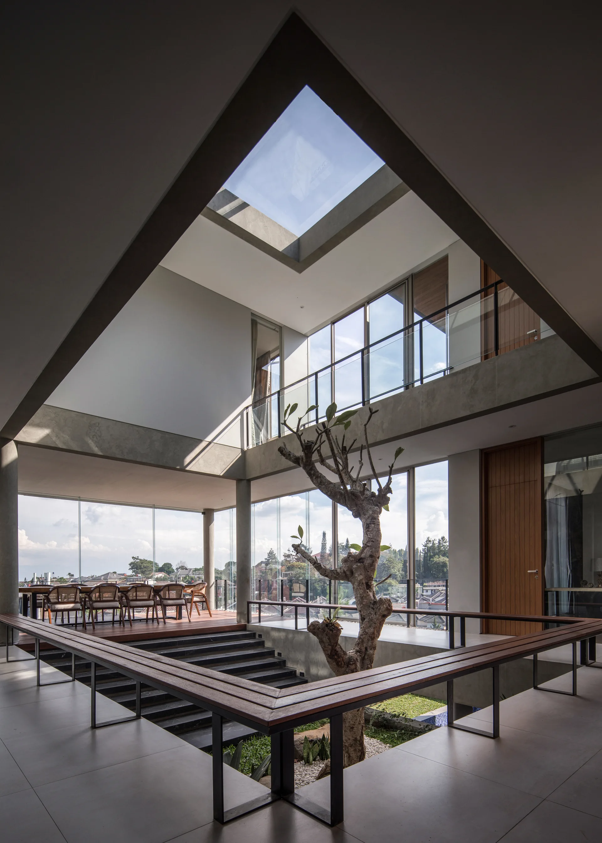 A modern indoor atrium featuring a sunken garden with a single tree, surrounded by wooden benches under a triangular skylight.