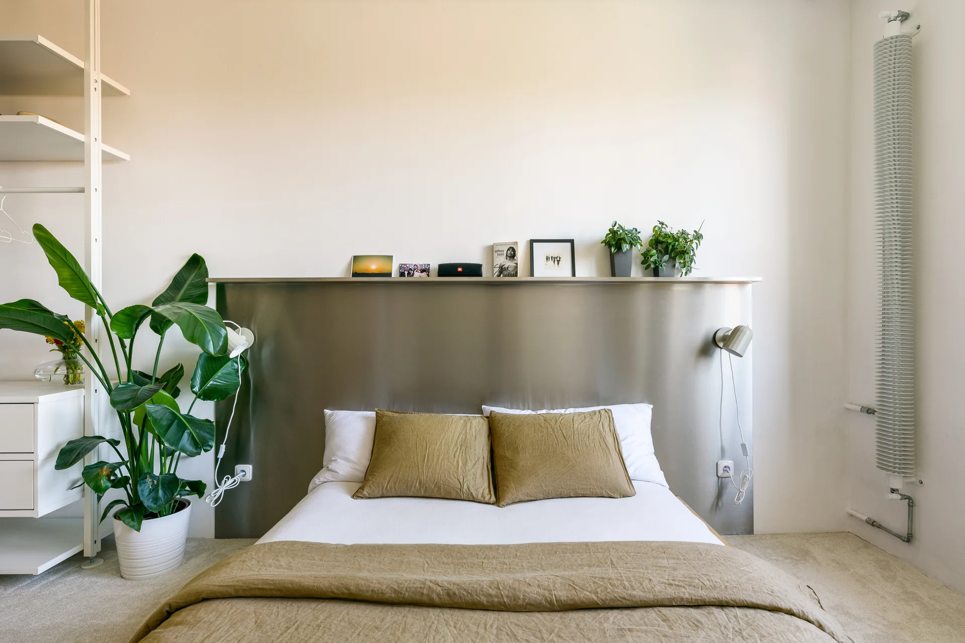A minimalist bed with olive green bedding sits against a brushed steel headboard panel featuring a narrow display ledge and sconces.
