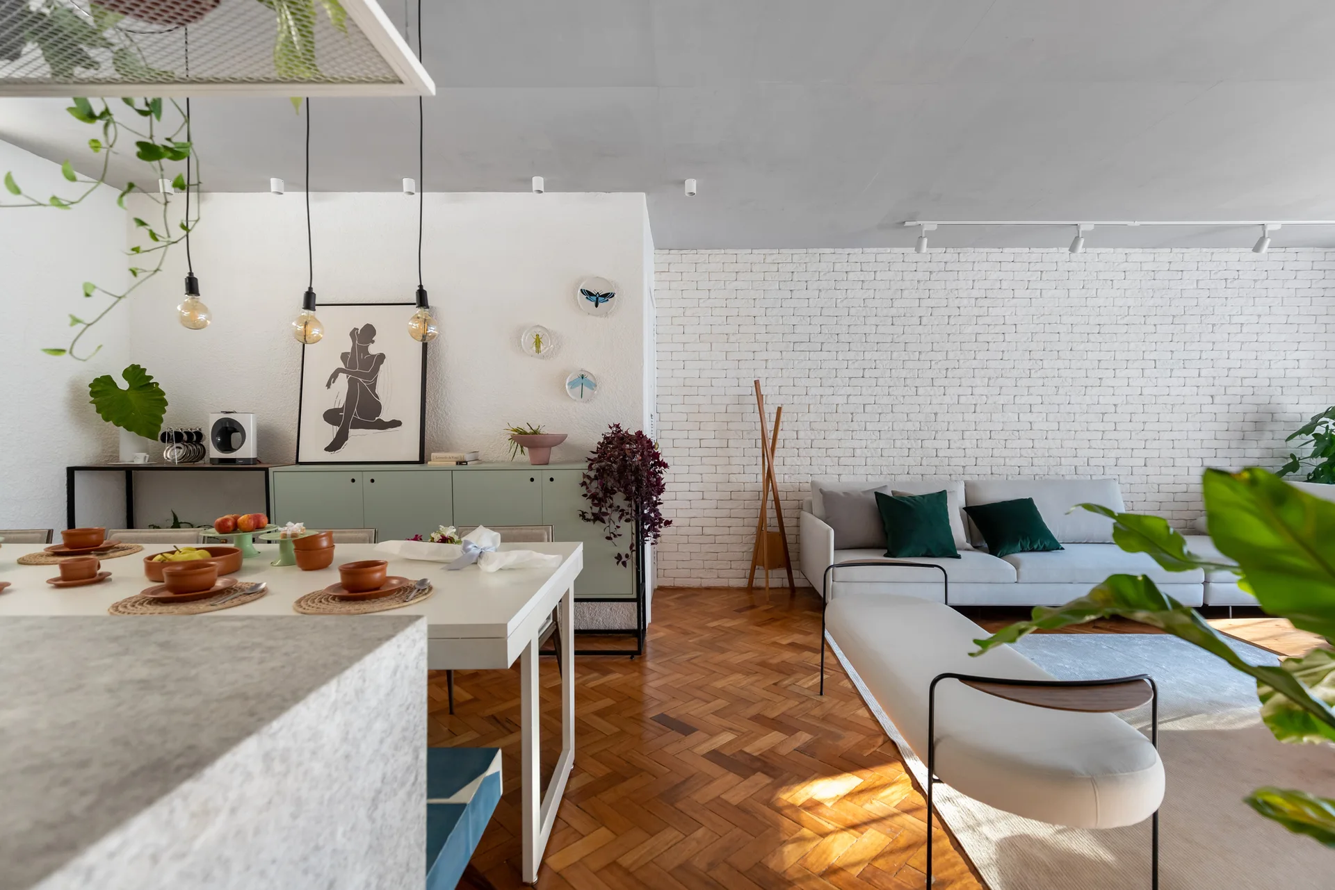 An open-concept living and dining space featuring a gray sofa, a white painted brick wall, a sage green sideboard, and herringbone wood floors.