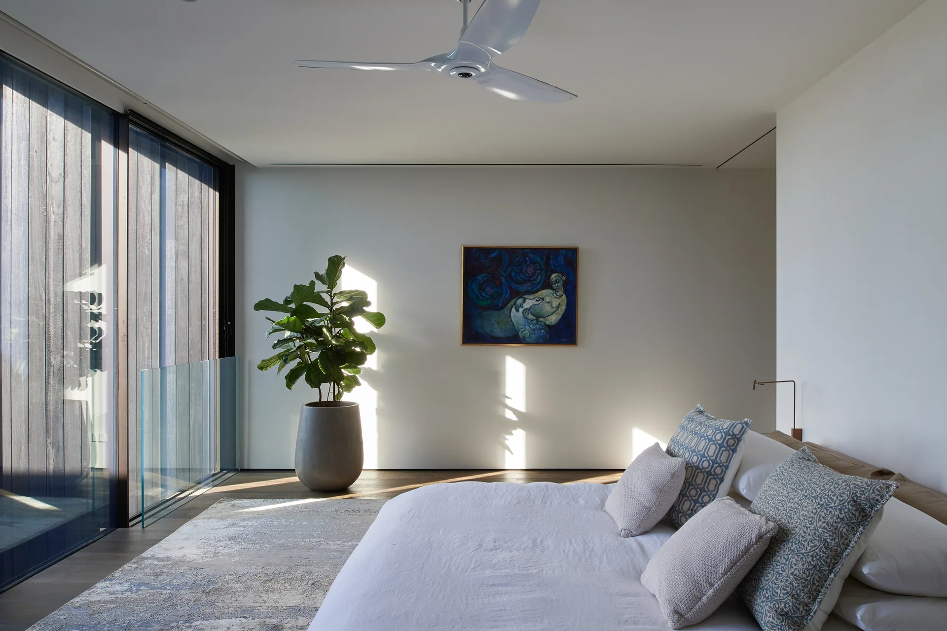 A minimalist white bedroom featuring a bed with patterned blue pillows, a large potted plant, and a sleek white ceiling fan.