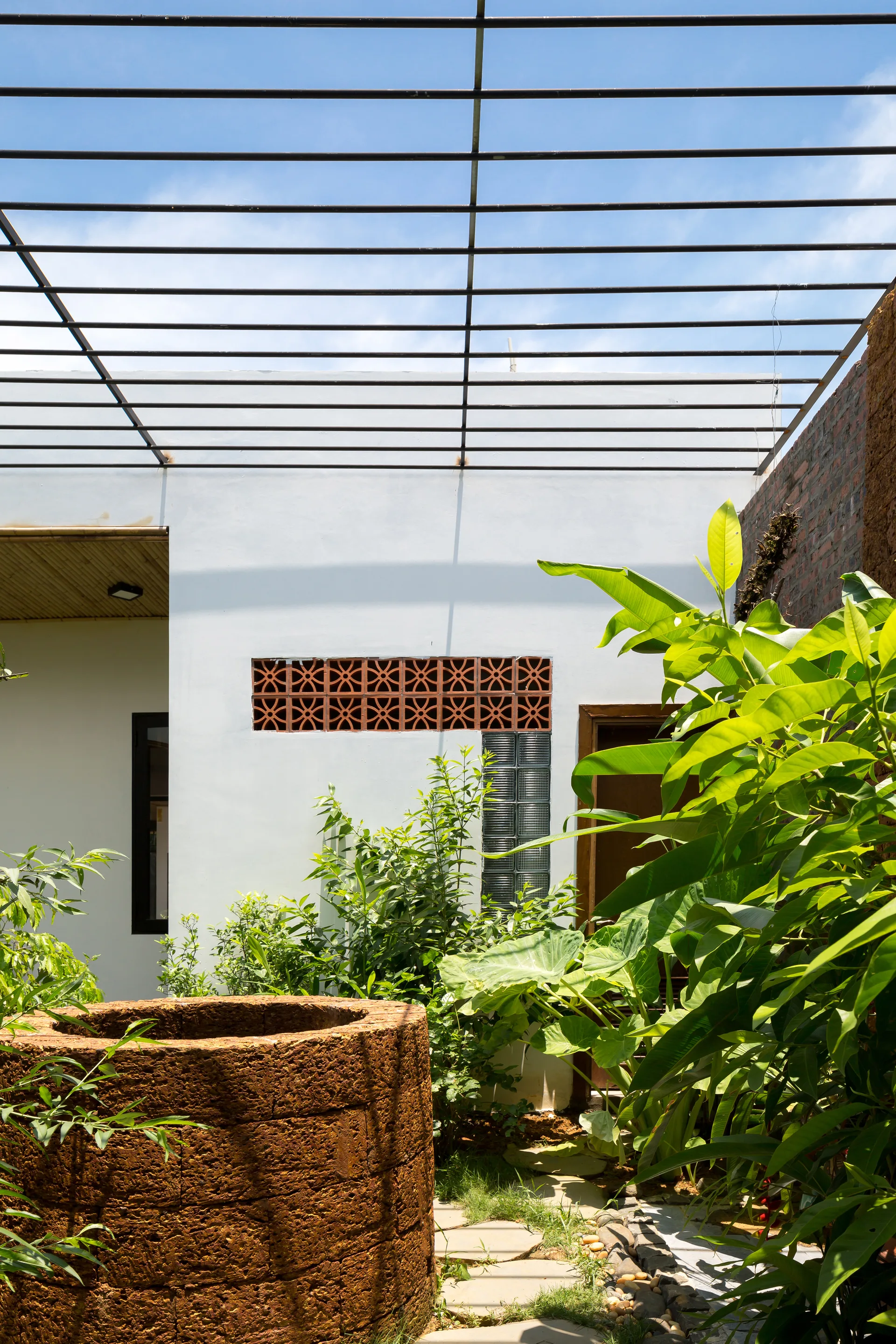 A sunny tropical courtyard featuring a circular stone well surrounded by lush green plants under a black metal slatted pergola.