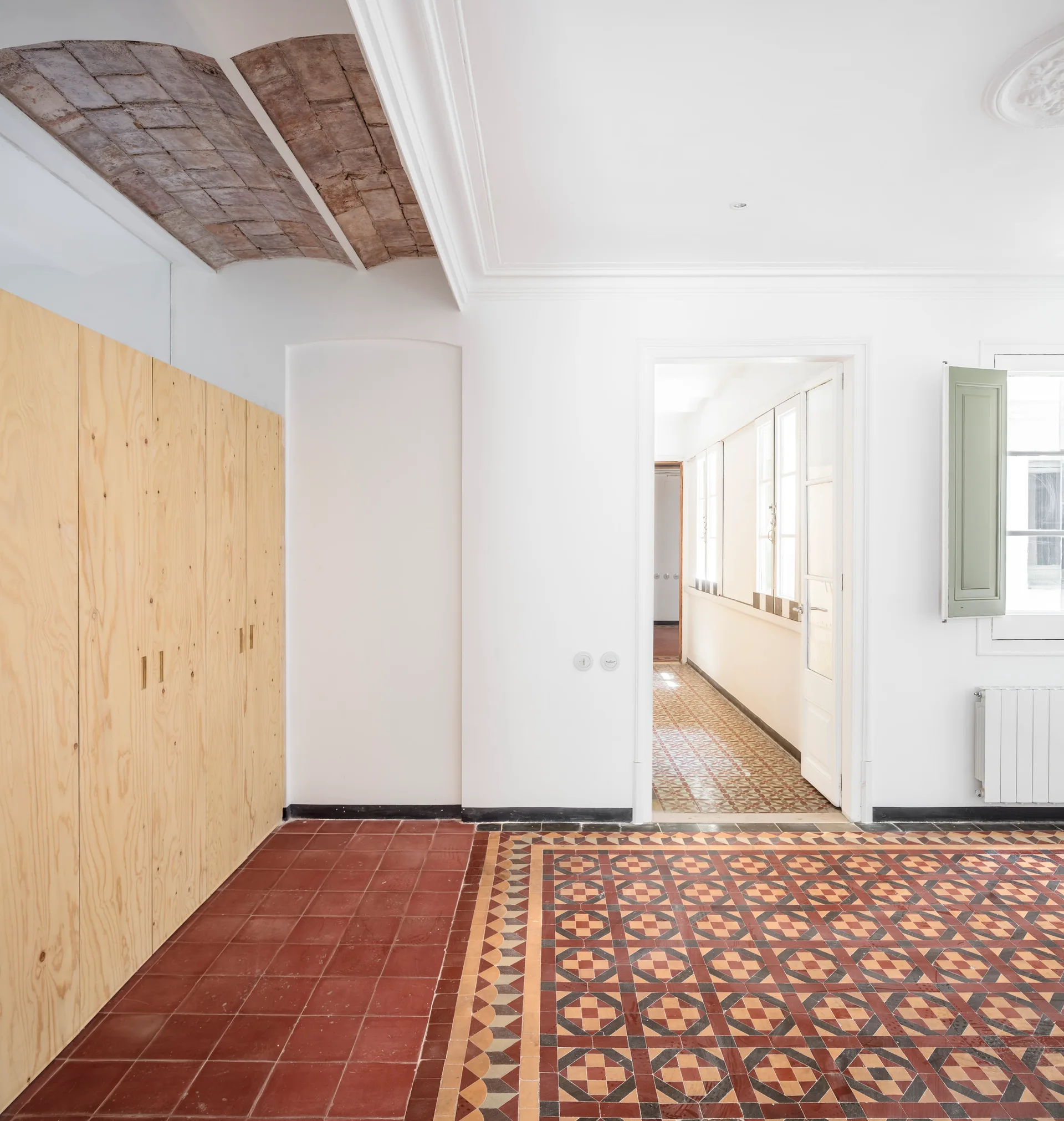 An empty room features patterned mosaic tile flooring, a custom light-wood built-in wardrobe, and an exposed brick archway ceiling detail.