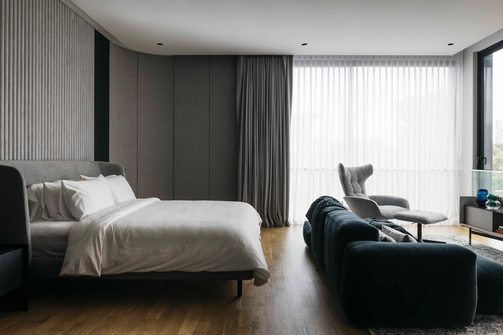 A contemporary bedroom features an upholstered bed, a dark blue sofa, and a grey wingback lounge chair near floor-to-ceiling drapes.