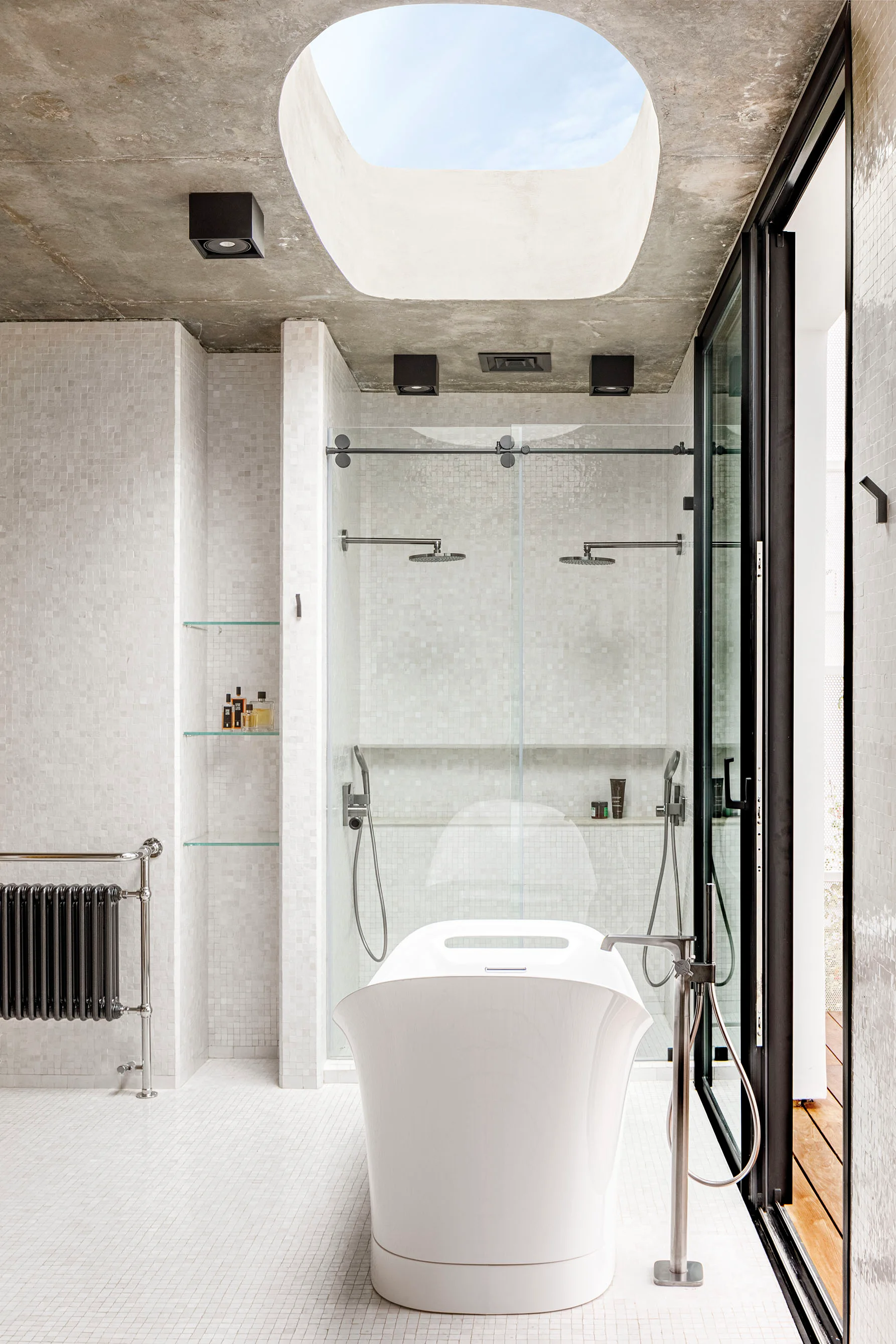 A modern bathroom features a white freestanding bathtub positioned beneath a large oval skylight in a raw concrete ceiling alongside white mosaic tiled walls.