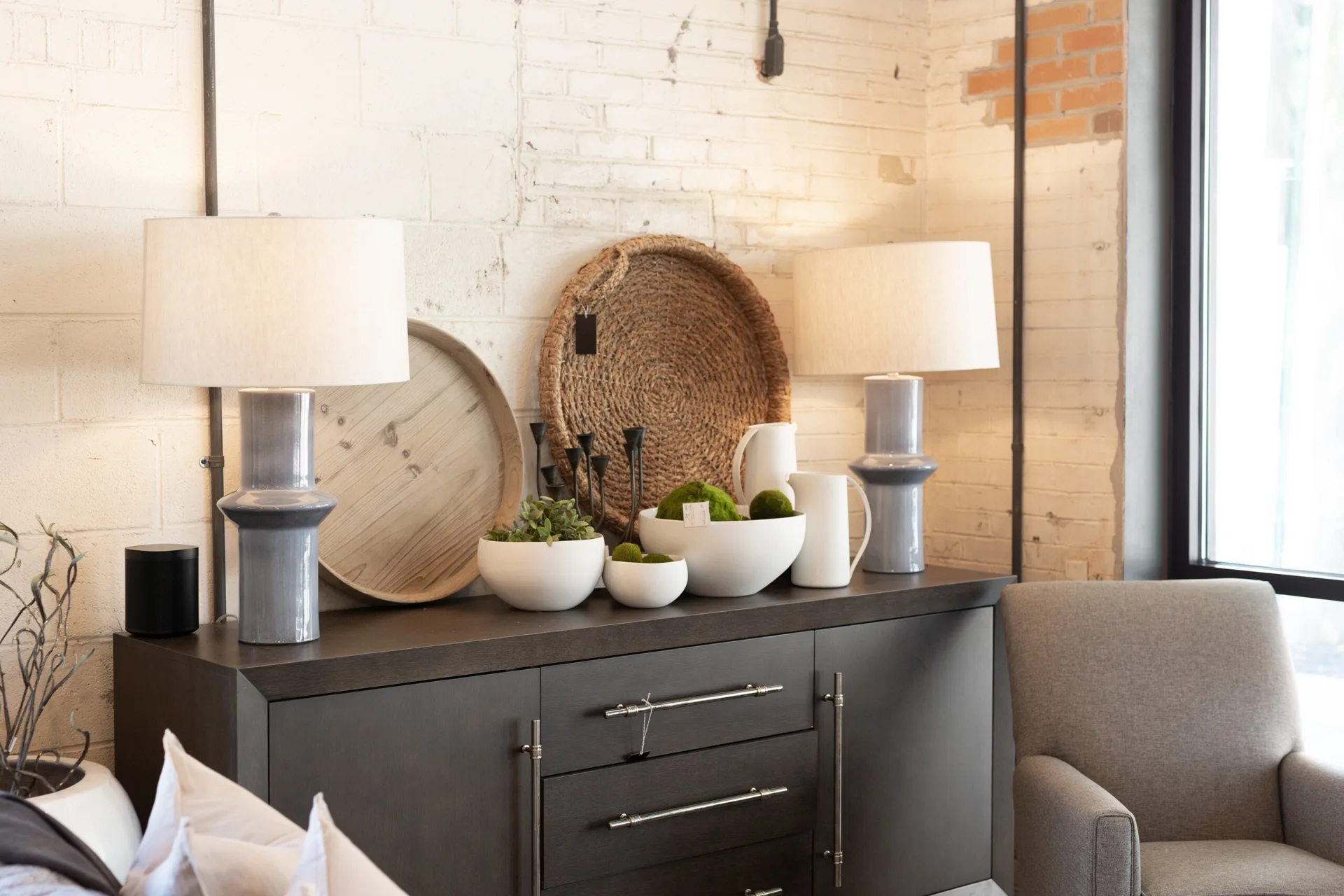 A dark wood buffet cabinet styled with gray ceramic lamps, white bowls, and woven trays against a painted white brick wall.
