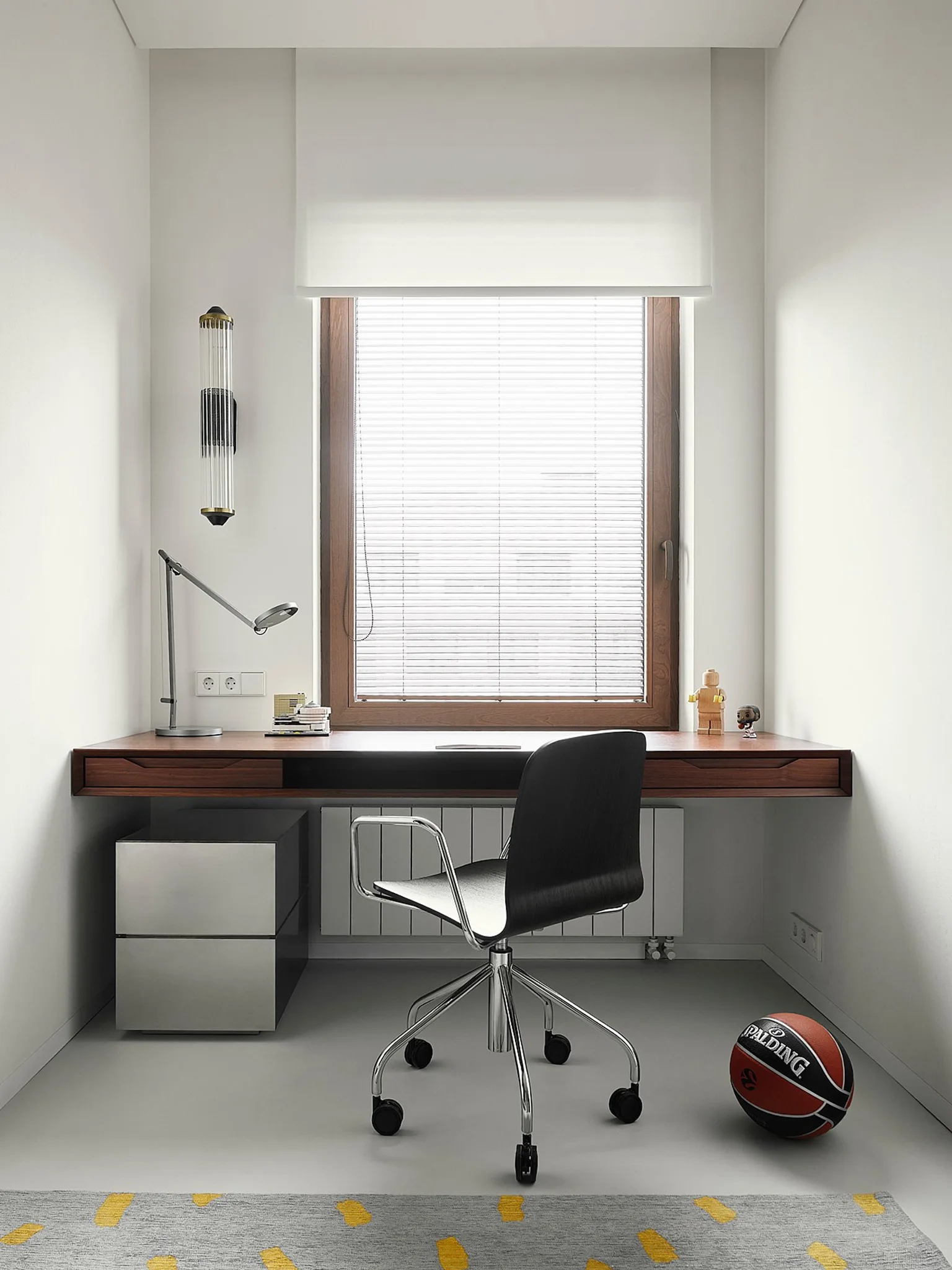 A minimalist home office alcove features a wall-mounted wooden desk beneath a window, a black rolling chair, and a metallic storage cabinet.