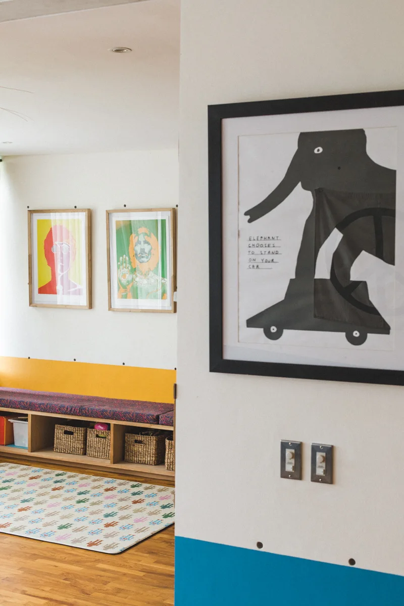 A bright residential hallway features a wooden storage bench with woven baskets, colorful framed artwork on the walls, and a patterned rug.