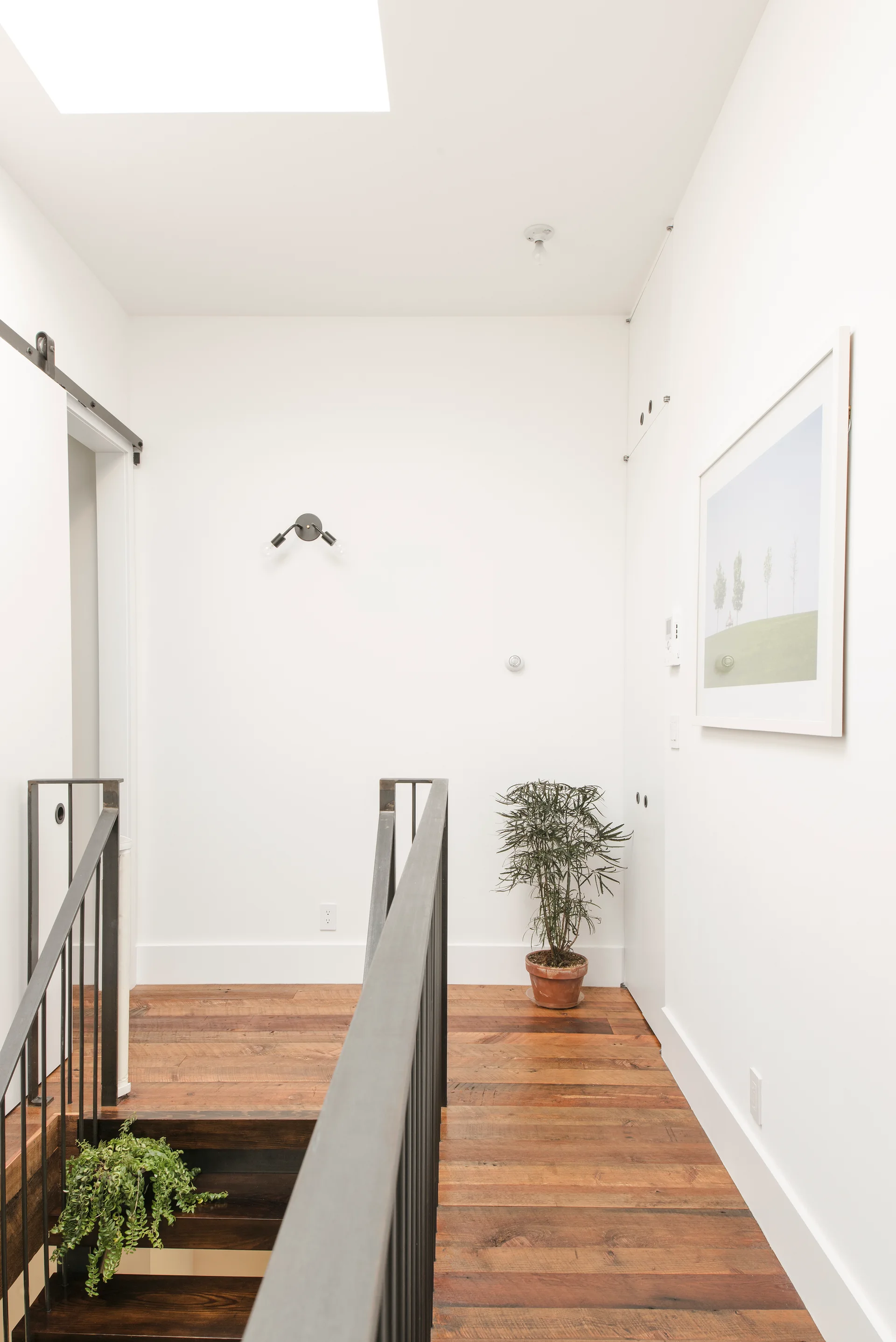 A minimalist staircase landing features wide-plank wood flooring, a black metal railing, and white walls illuminated by natural light from a skylight.