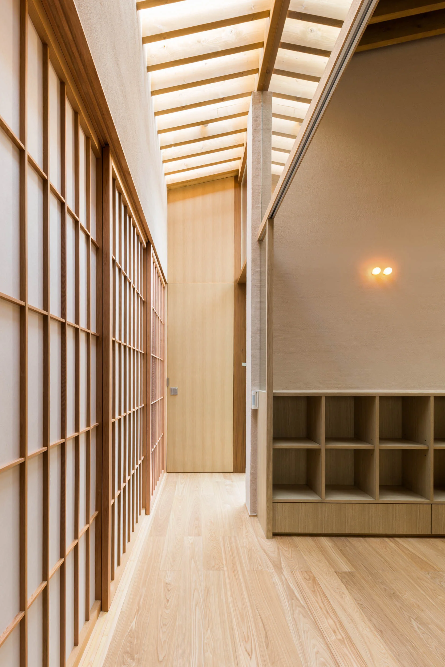 A minimalist hallway featuring wooden shoji screens, a slatted timber ceiling with skylight, light wood floors, and low open cubby shelving.