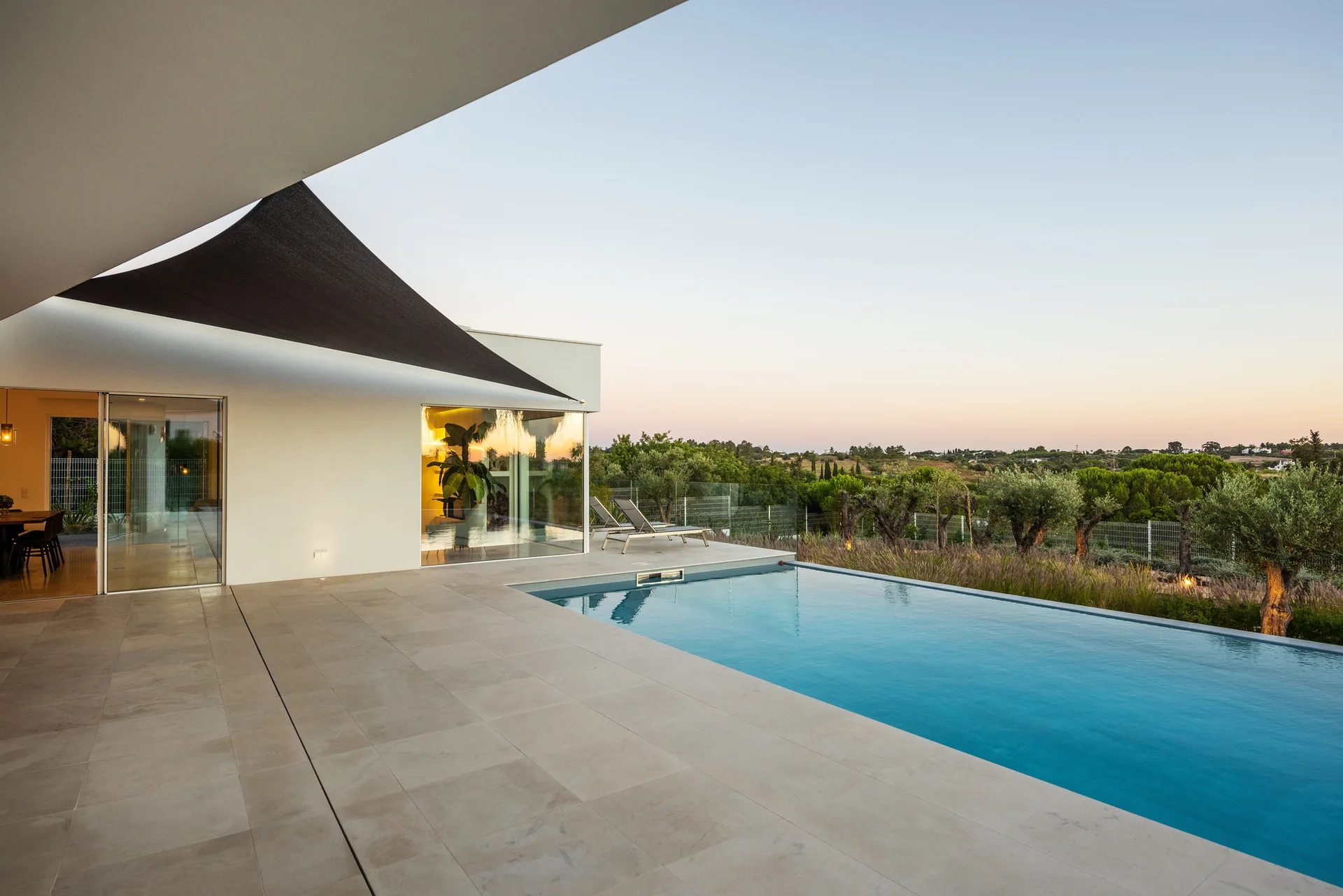 A rectangular swimming pool is flanked by a large-format beige tiled patio, a white modern house with floor-to-ceiling glass, and a black shade sail.