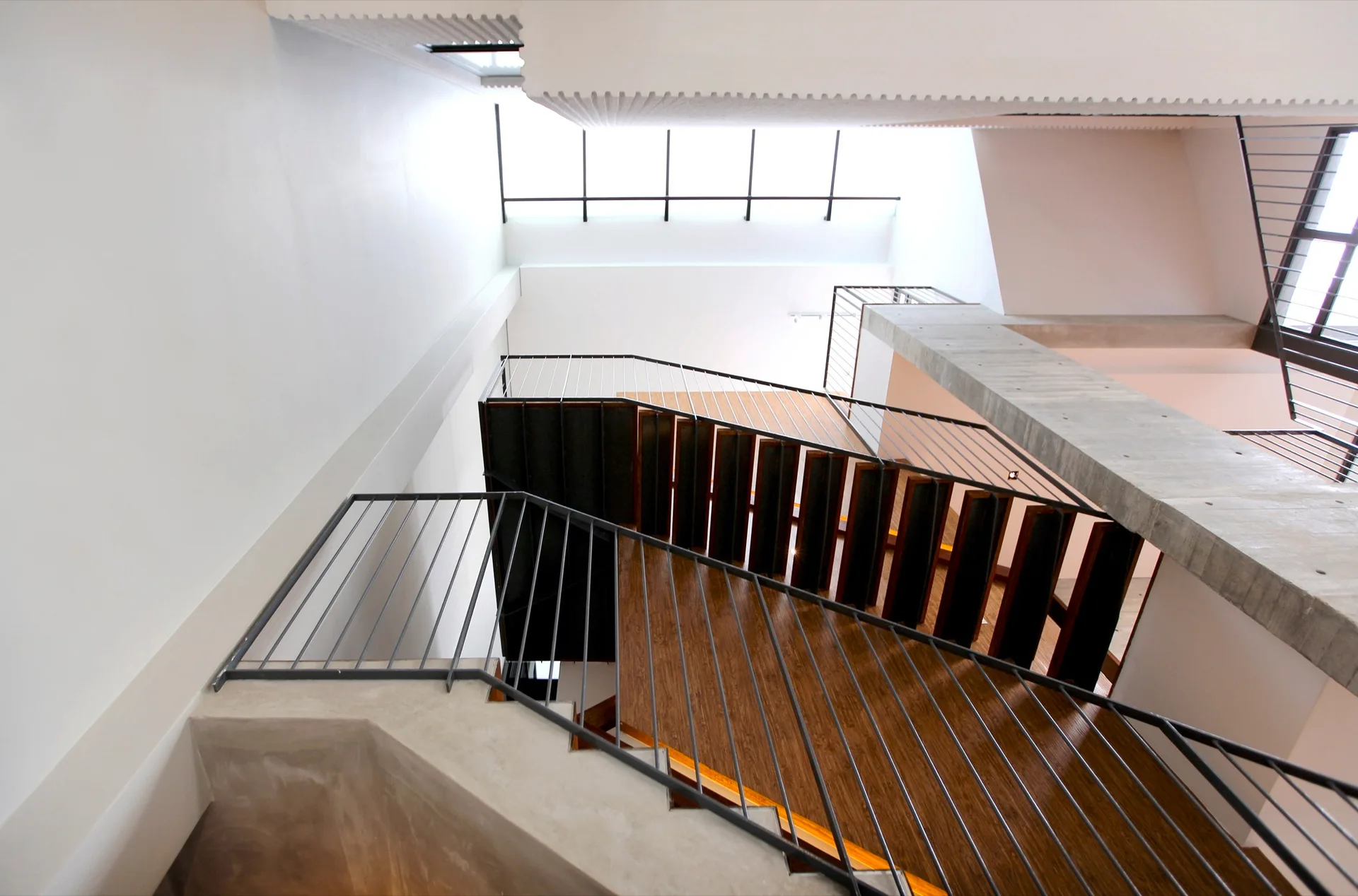 Looking up a modern multi-level staircase featuring warm wood treads, slender black metal railings, and a bright overhead skylight.