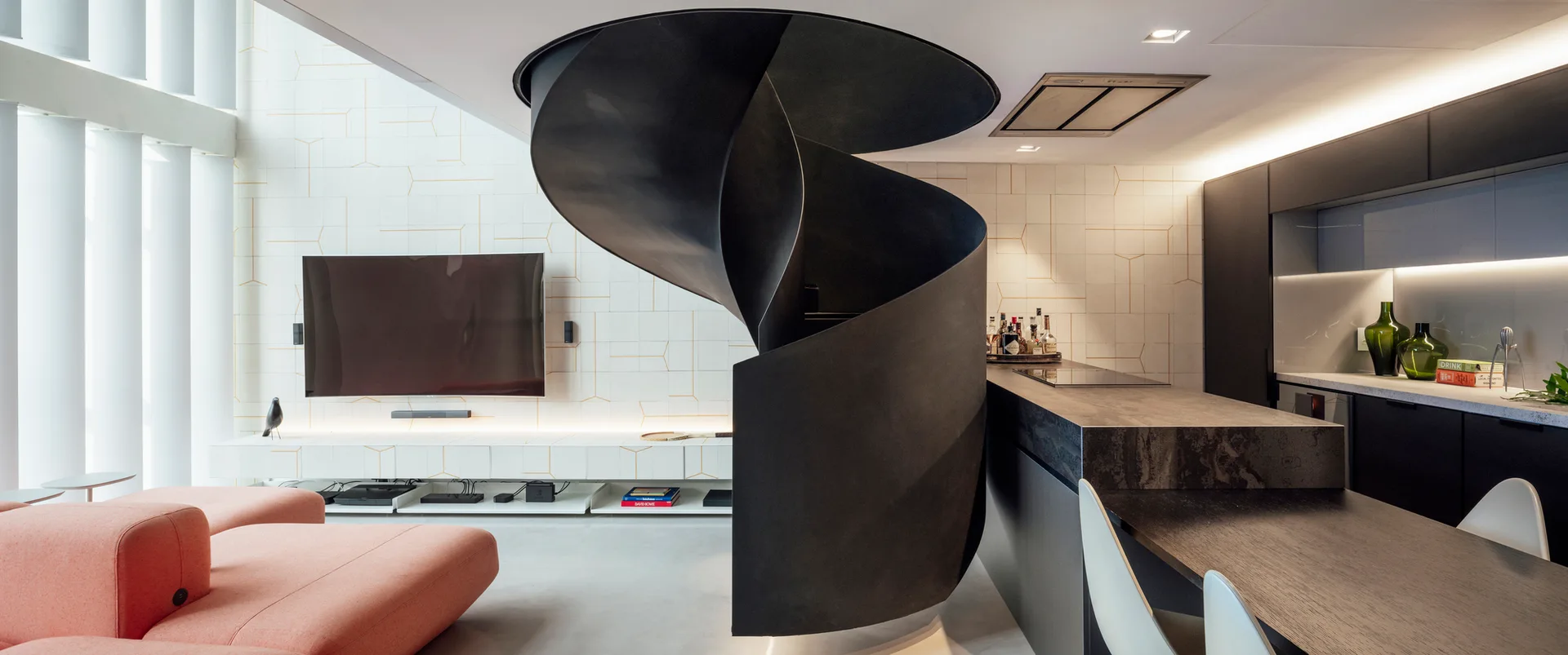 A modern open-plan living room and kitchen featuring a central black spiral staircase, a salmon-colored sectional sofa, and a geometric-patterned wall.