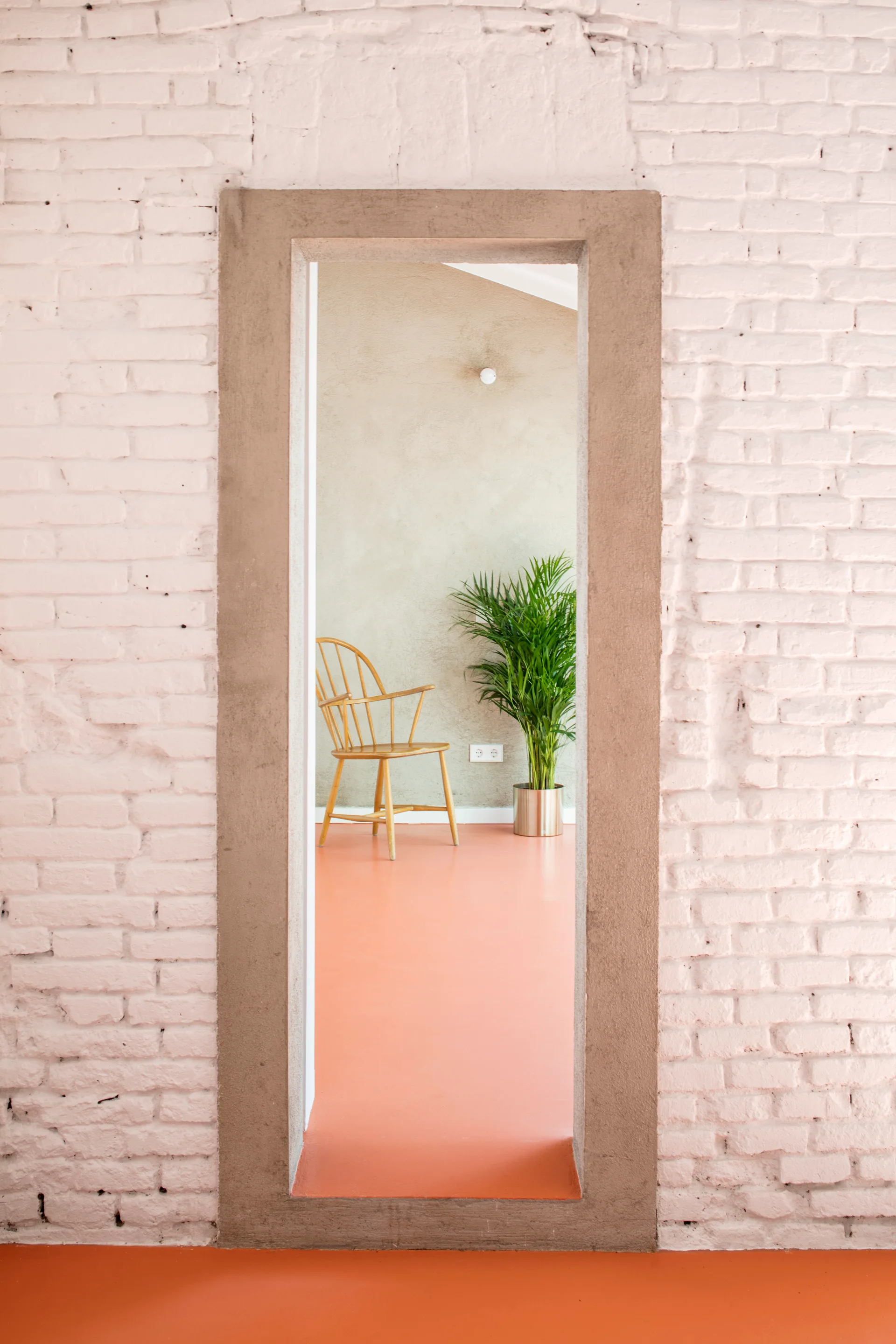 A view through a stone doorway in a white brick wall shows a minimalist room with a bright orange floor, a wooden chair, and a green plant.