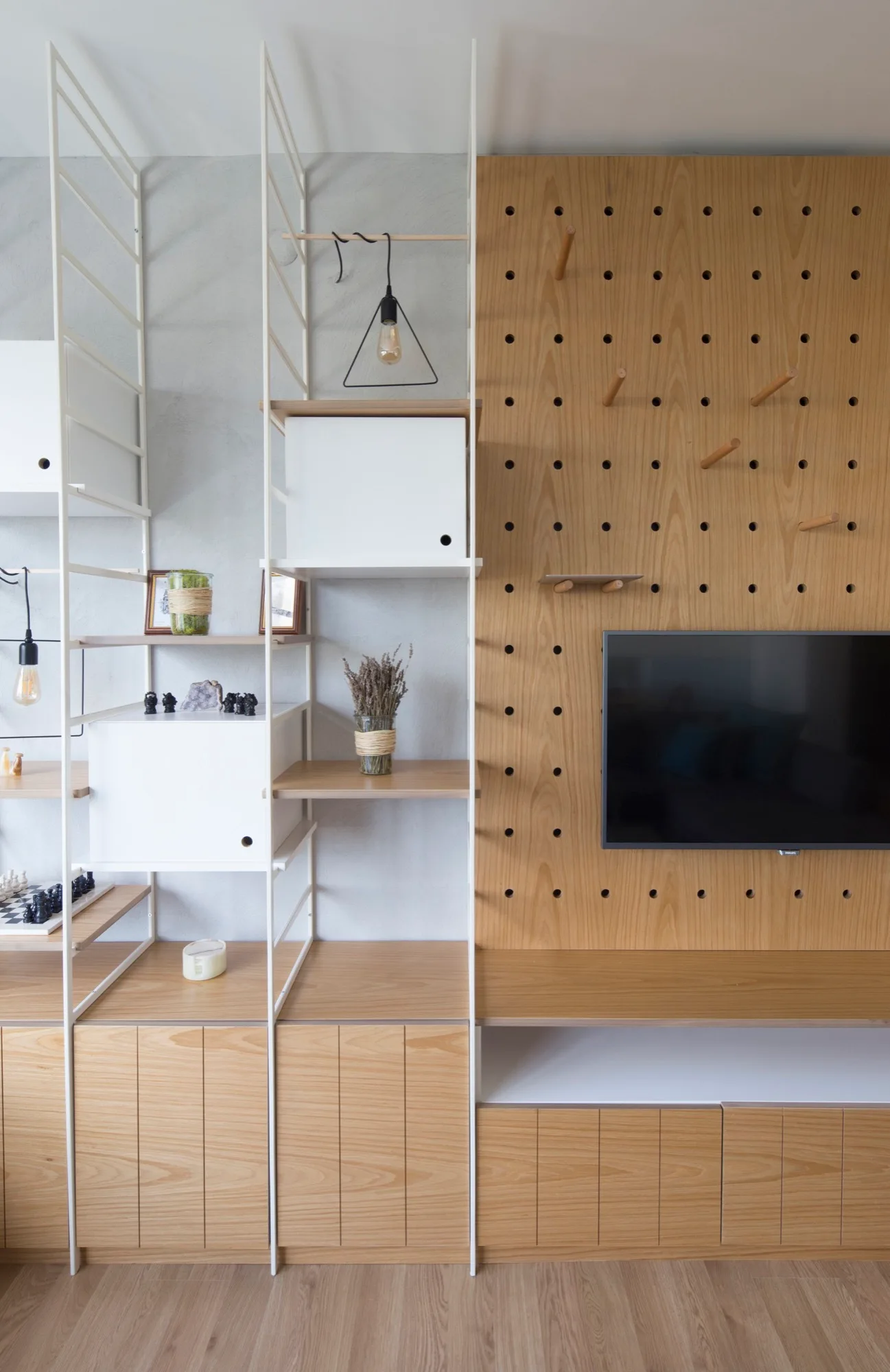 A modern wooden pegboard wall panel with a mounted TV next to white metal modular ladder shelving and wood cabinets.