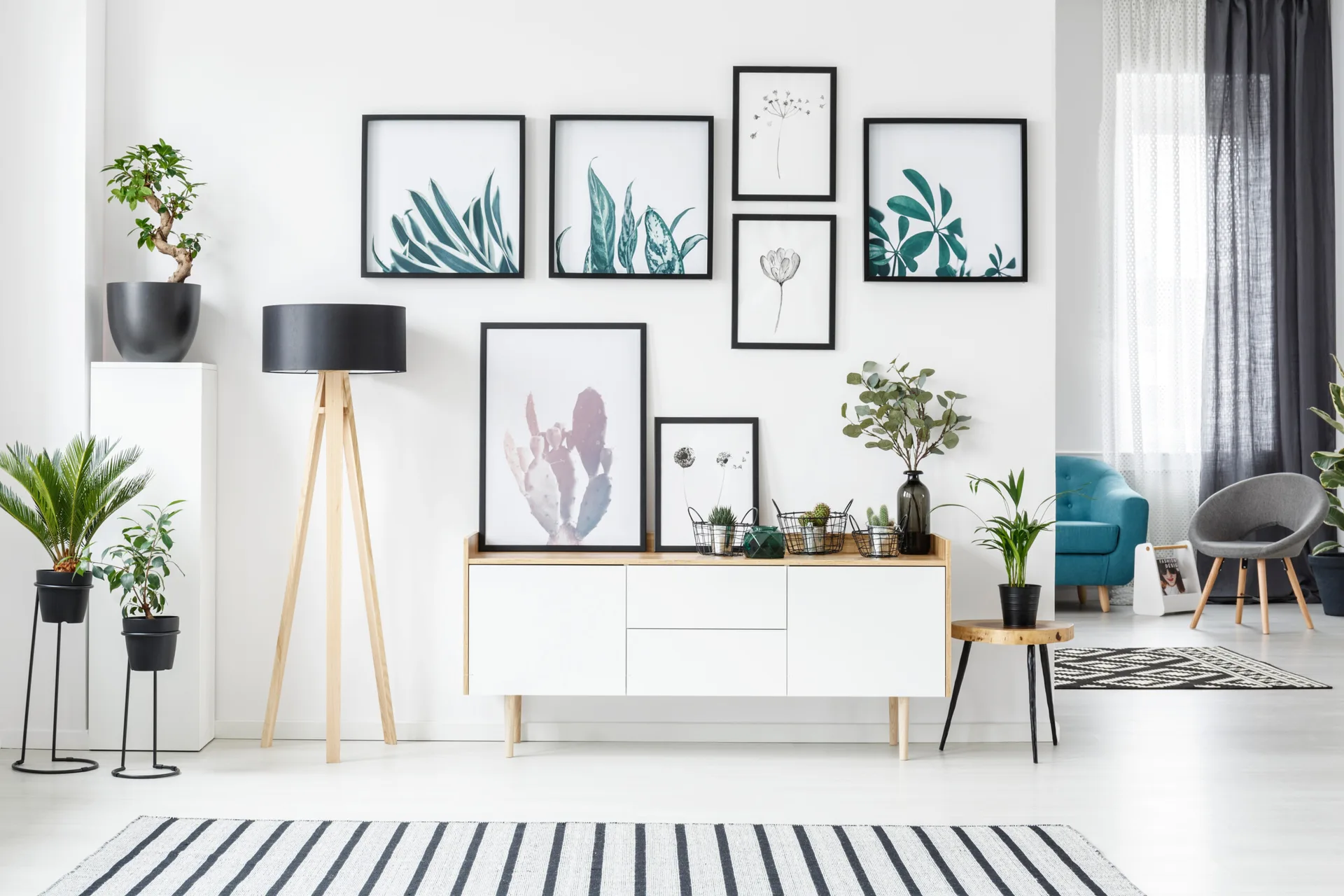 A white Scandinavian console table stands beneath a botanical gallery wall, flanked by a wooden tripod lamp and green potted plants.