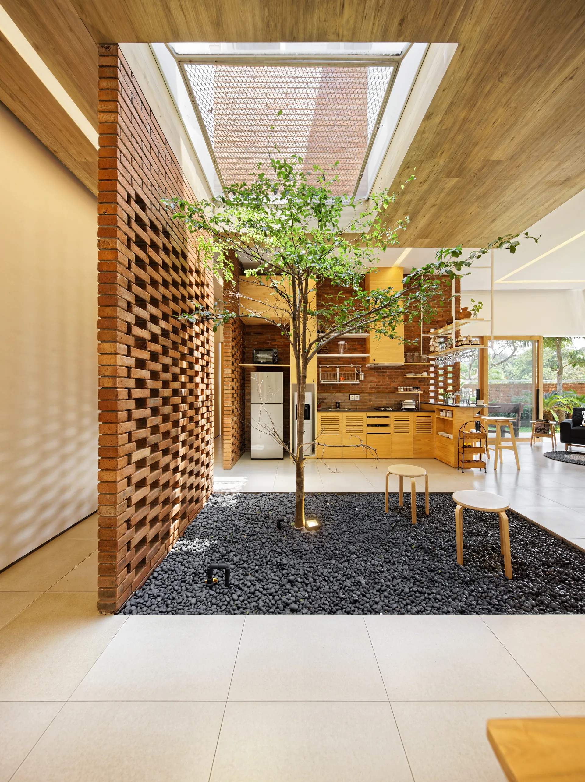 An indoor garden with a tree planted in black pebbles sits beneath a skylight next to an open kitchen with wood cabinets and a brick screen wall.