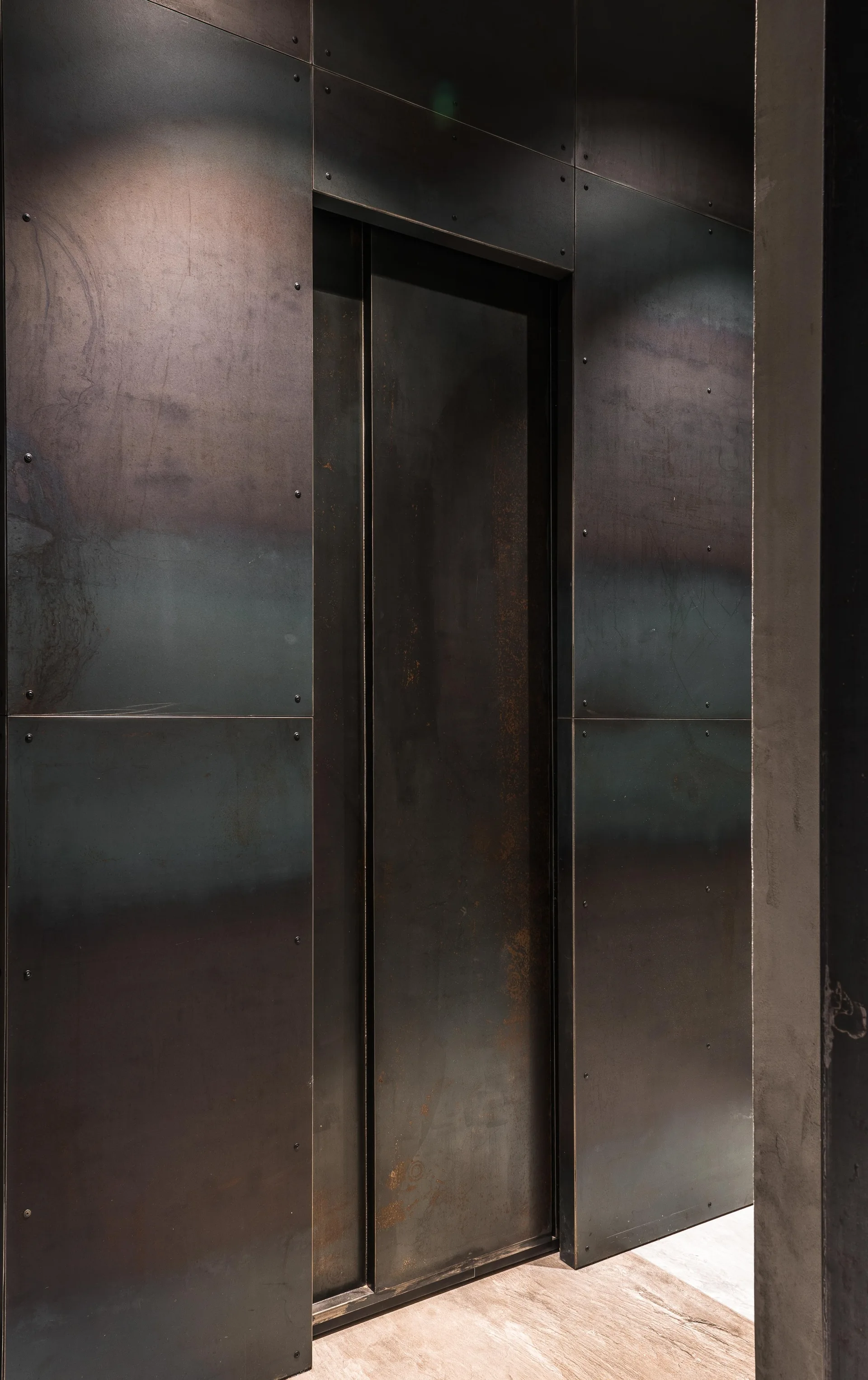 Dark metal panels with exposed rivets line the walls of an industrial-style hallway surrounding a recessed charcoal metal door.