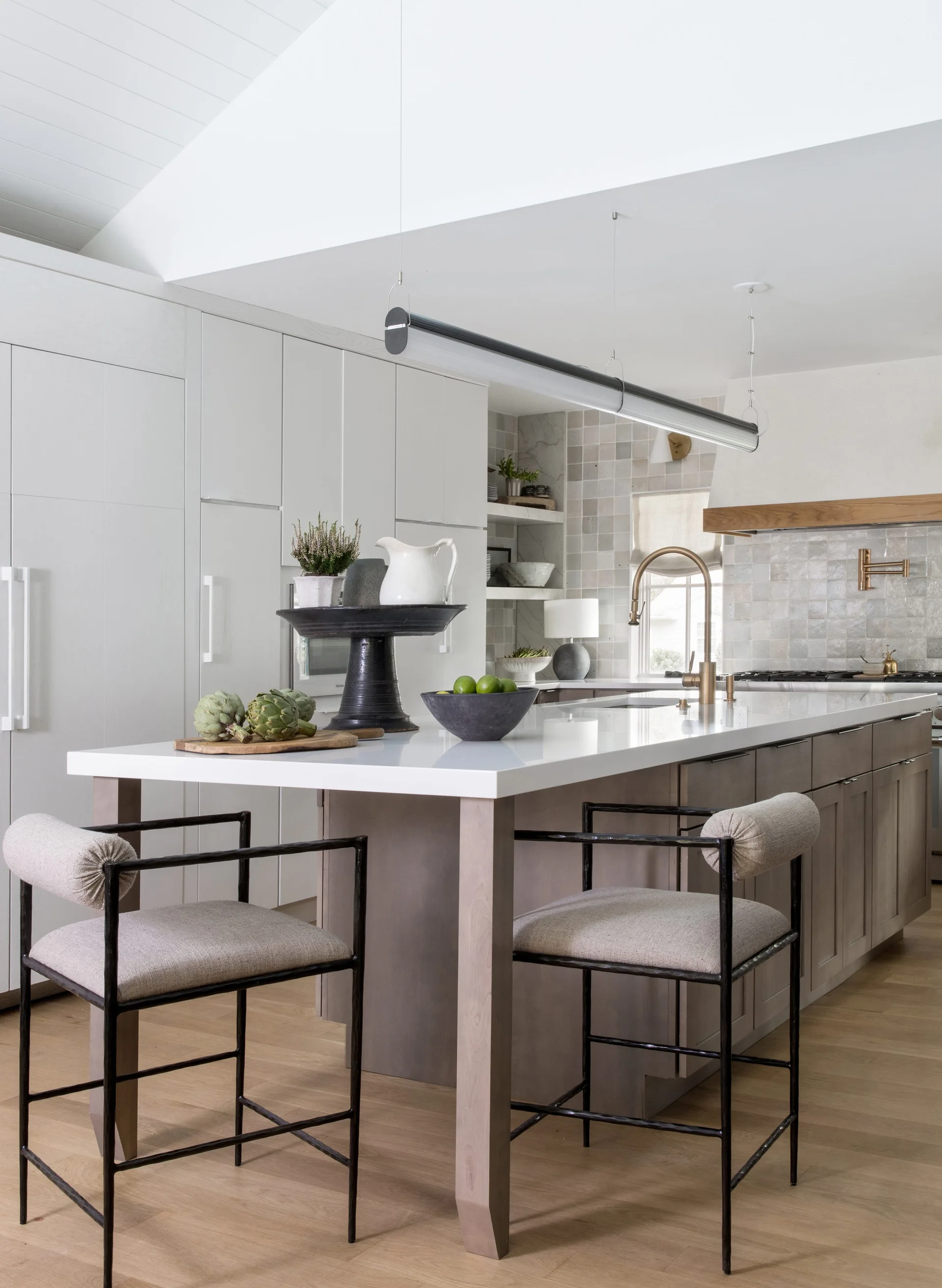 A modern kitchen featuring a light grey island with upholstered stools, a cylinder pendant light, and a square tile backsplash.