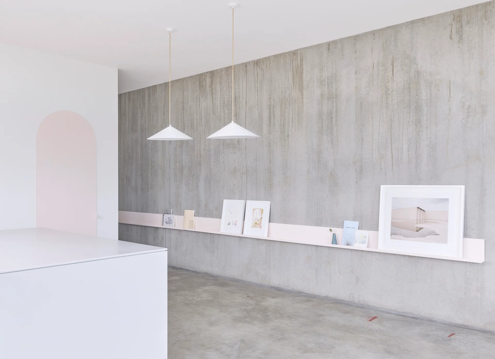 A textured concrete wall features a long pale pink display ledge holding books and picture frames under two white conical pendant lights.