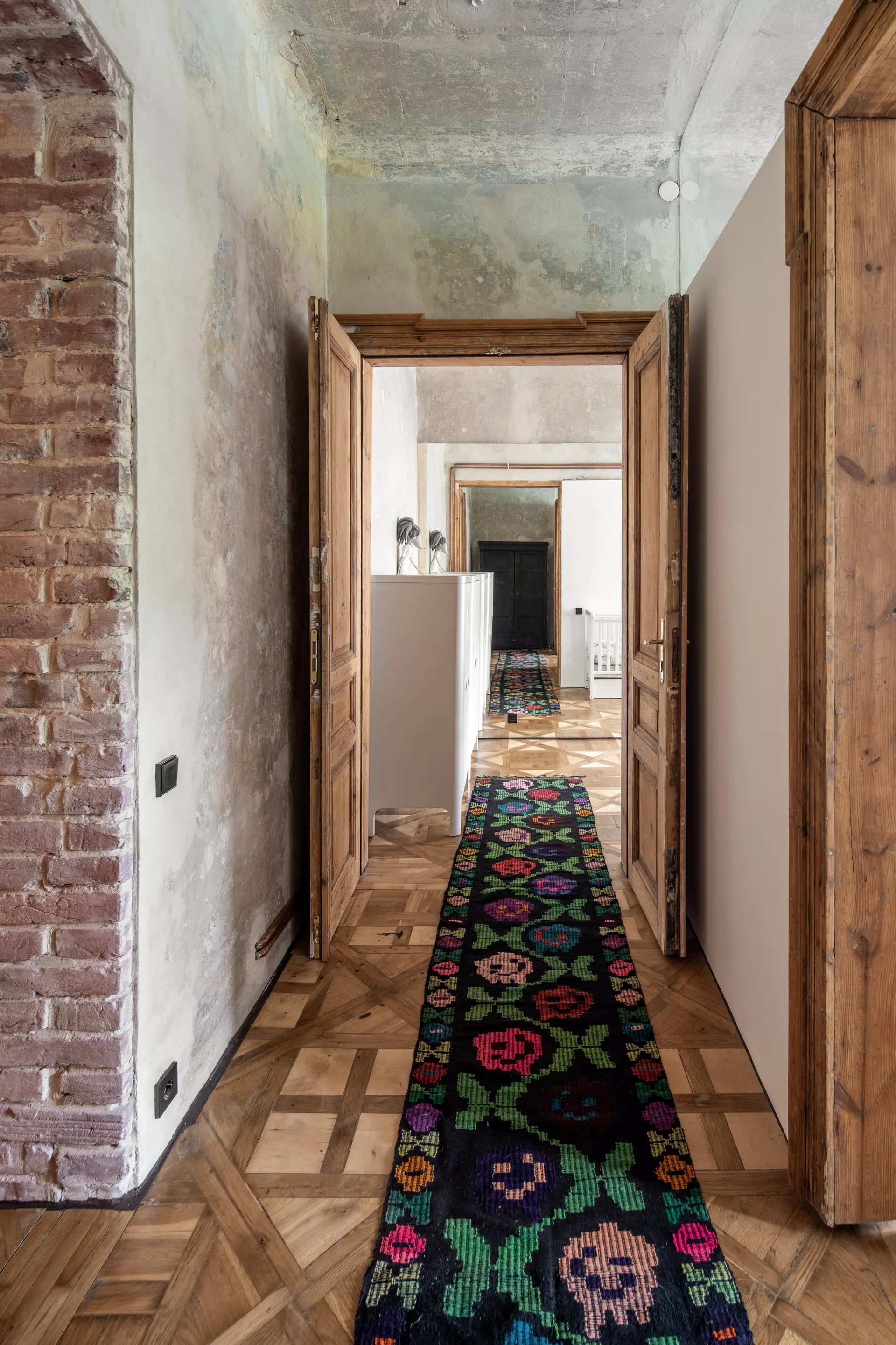 A perspective view of connected rooms through wooden doorways with parquet flooring, exposed brick, and a long black floral runner rug.