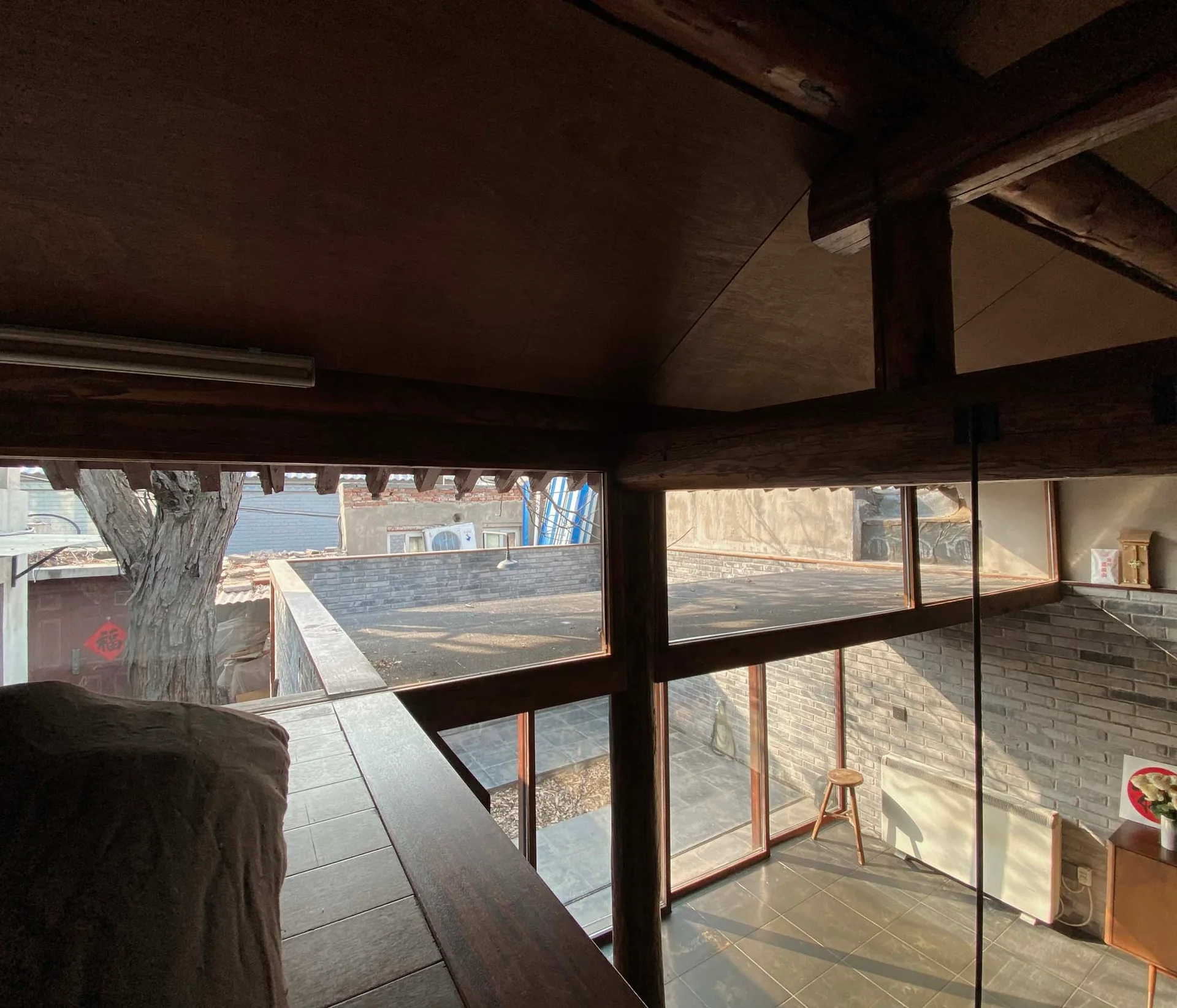 View from an upper loft level looking down at a tiled room with grey brick walls, wood beams, and large windows opening to a courtyard.